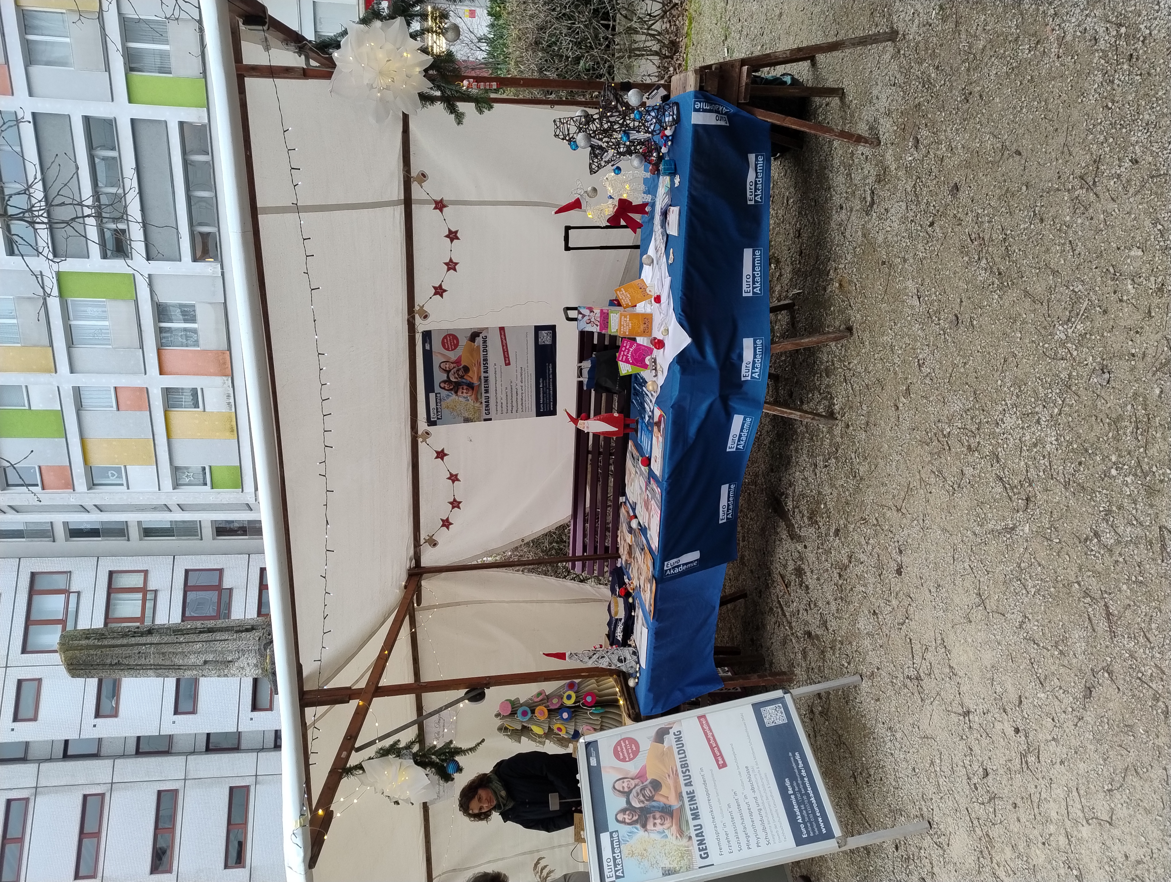 Überdachter Infostand der Euro Akademie auf einem Platz zwischen Hochhäusern; auf dem blau gedeckten Tisch liegen Flyer und kleine Bastelarbeiten, im Hintergrund hängen Lichterketten und ein großes Plakat „Genau meine Ausbildung“.