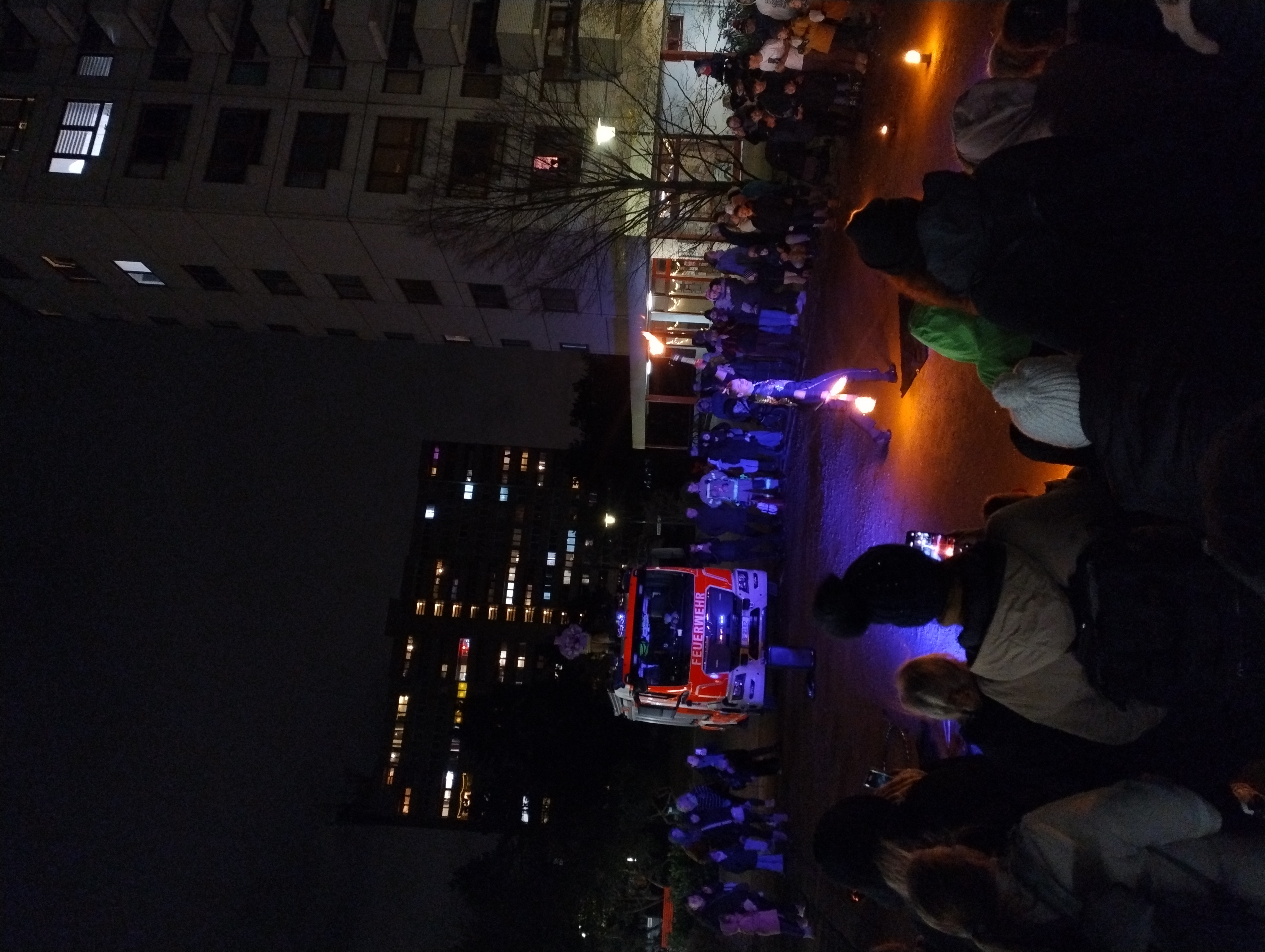 Große Menschenmenge schaut im Dunkeln einer Feuershow zu; vor einem Feuerwehrfahrzeug mit der Aufschrift „Feuerwehr“ jongliert eine Artistin mit brennenden Fackeln, ringsum hohe Wohnhäuser mit erleuchteten Fenstern.
