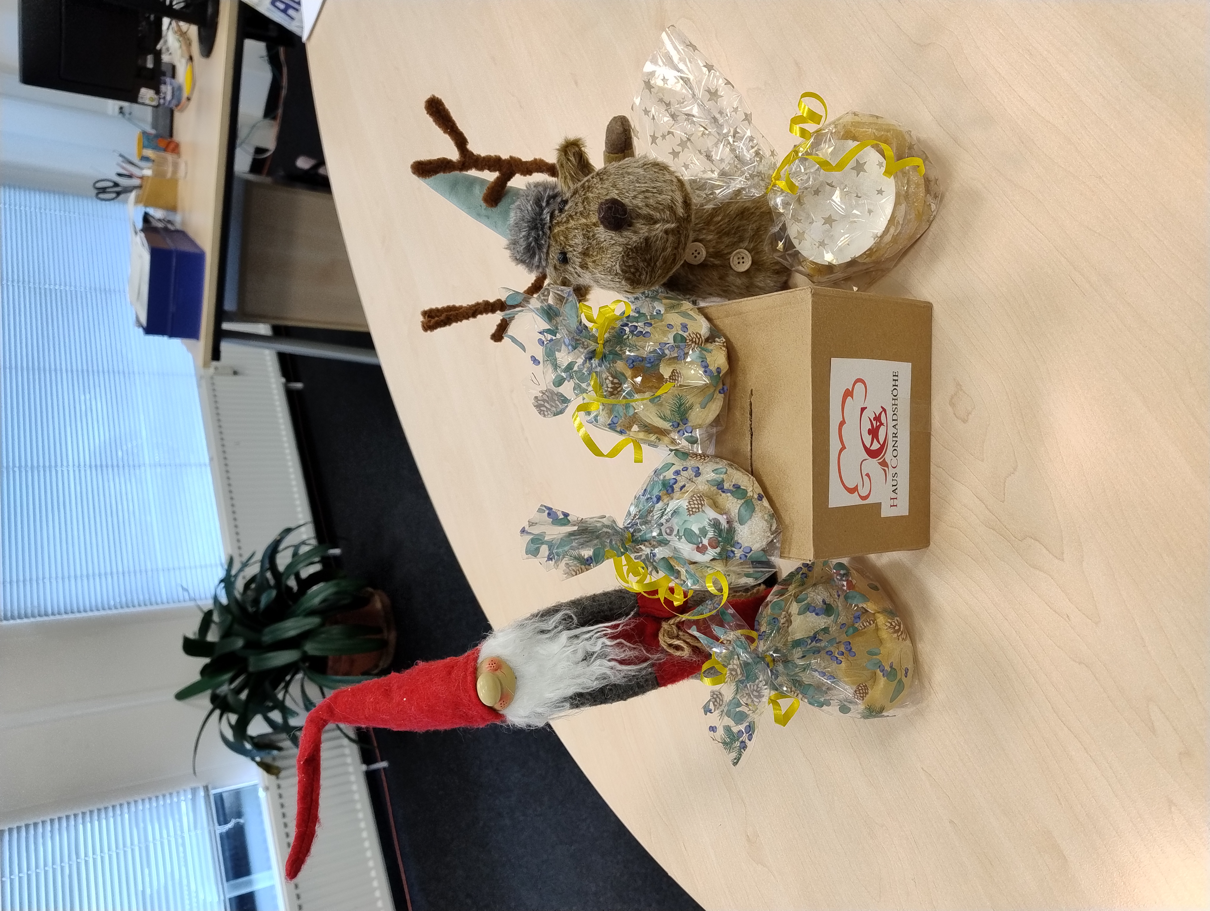 Auf einem Schreibtisch stehen ein Weihnachtswichtel und ein Plüschrentier neben einer Spendenbox „Haus Conradshöhe“, umgeben von mehreren durchsichtigen Geschenkbeuteln mit gelben Schleifen.