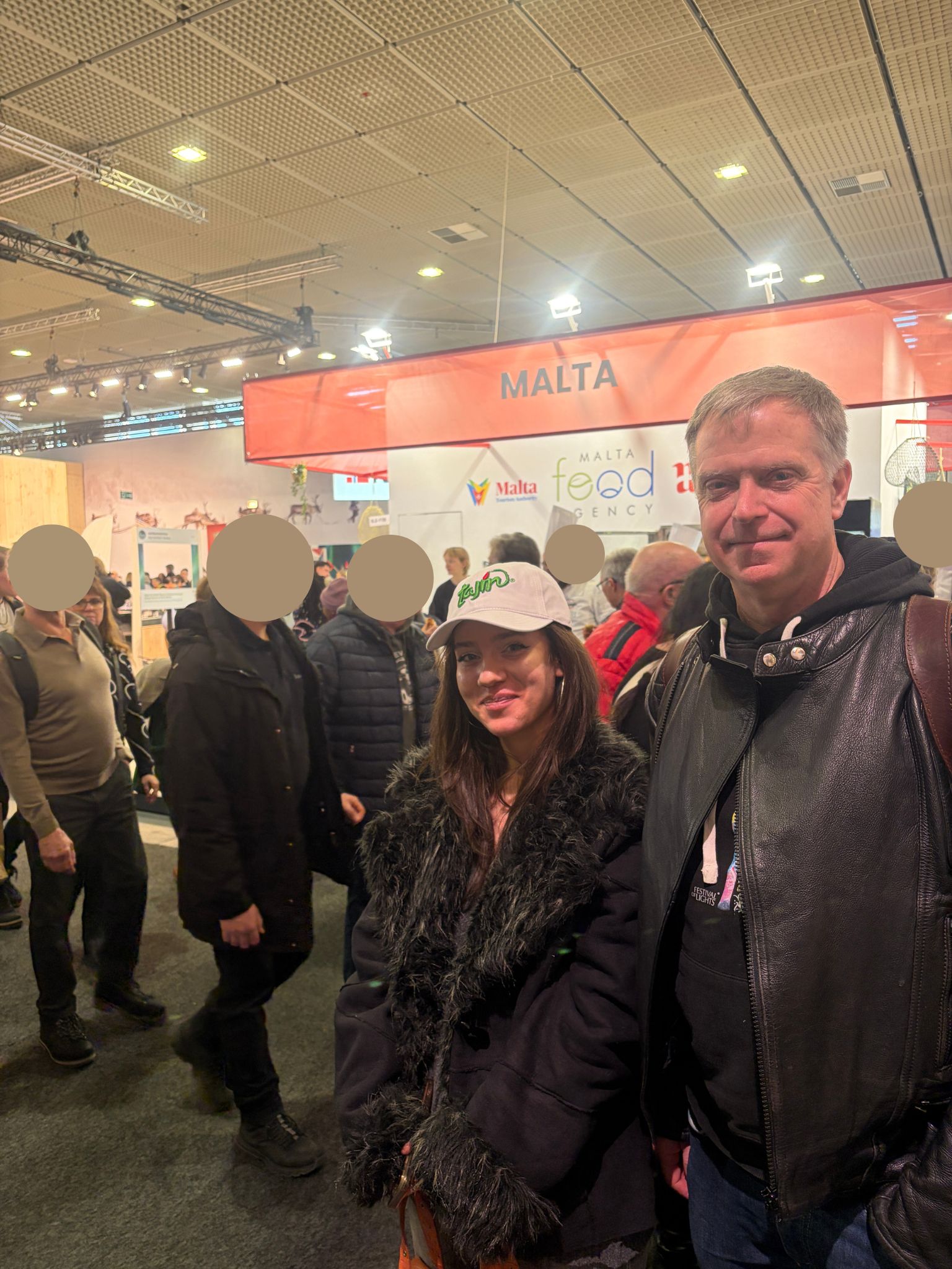Schülerin und Lehrer in einer Messehalle voller Leute, im Hintergrund ein Banner mit der Aufschrift "Malta"