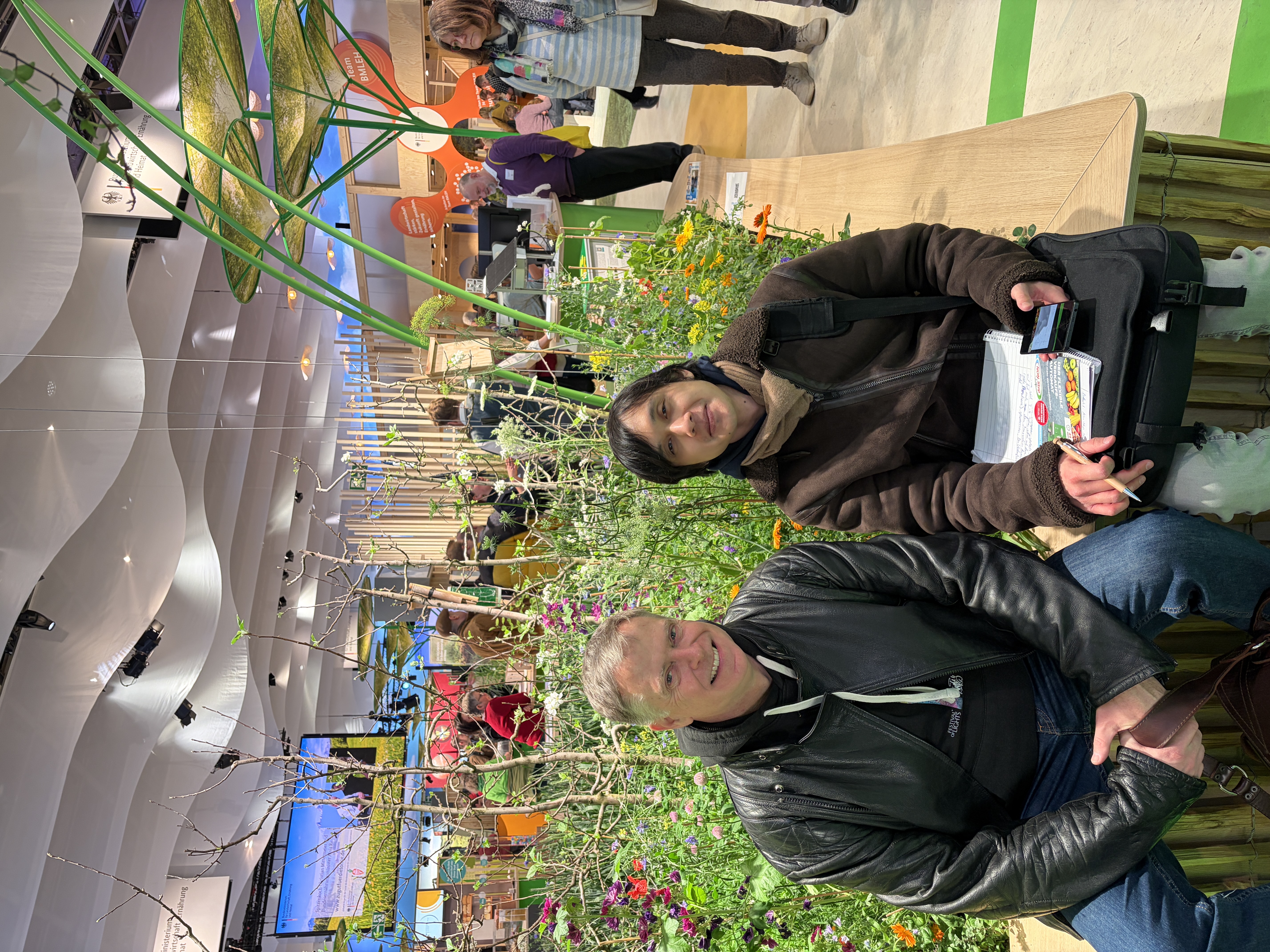 Lehrer und Schüler sitzen vor einem bunten Blumenbeet in einer Messehalle