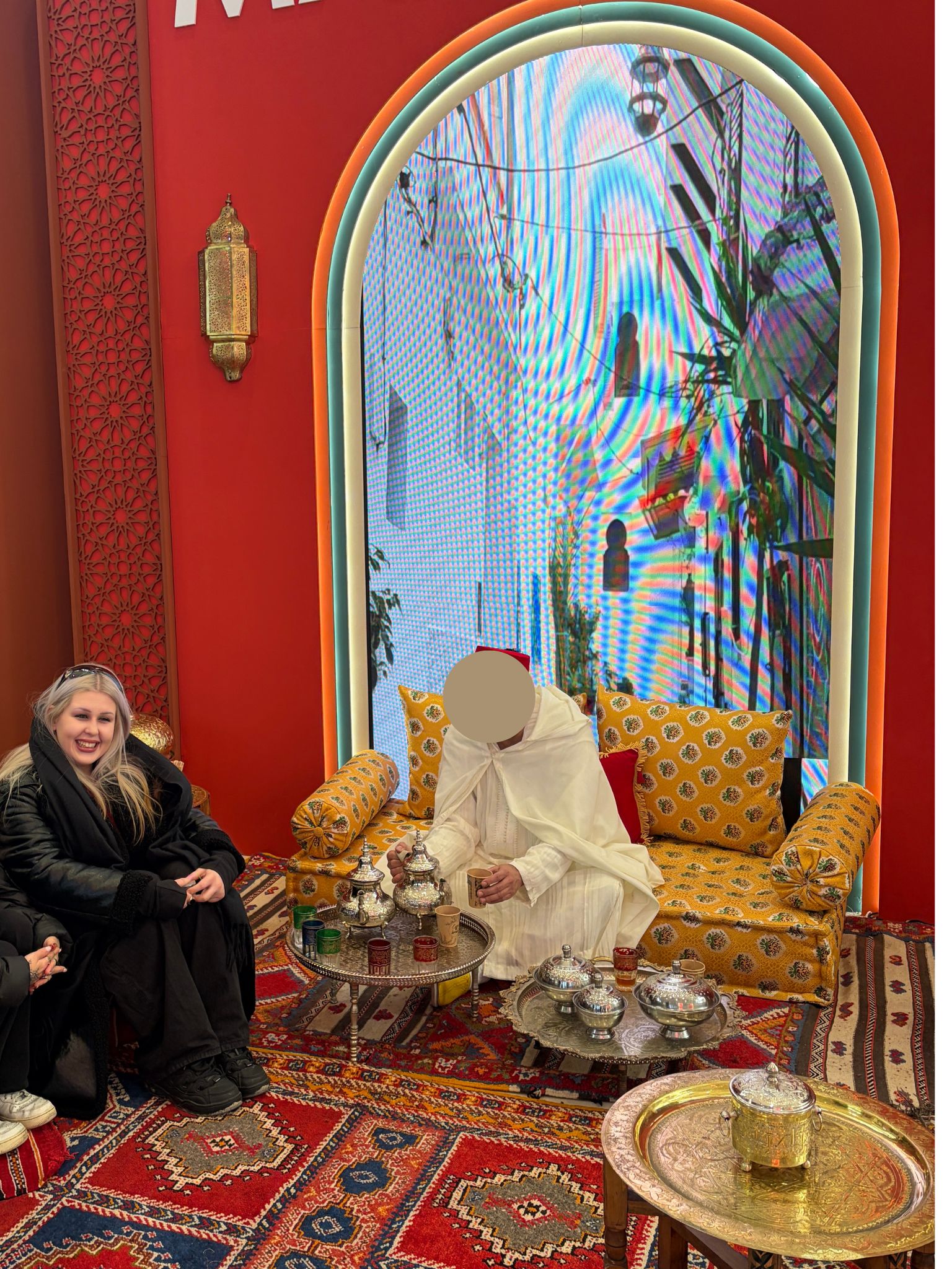 Schülerin sitzt lächelnd bei einer marokkanischen Teezeremonie; ein Mann in traditioneller Kleidung sitzt auf einem gelben Sofa und schenkt Tee aus; auf dem Boden und an der Wand sind rote Teppiche