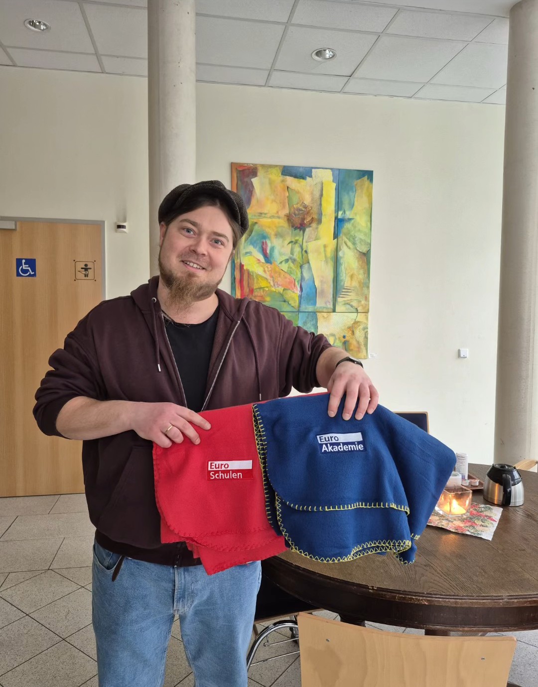 Eine Person steht in einem hellen Innenraum neben einem runden Tisch und hält zwei zusammengefaltete Decken in die Kamera: eine rote mit dem Logo ‚Euro-Schulen‘ und eine blaue mit dem Logo ‚Euro Akademie‘.
