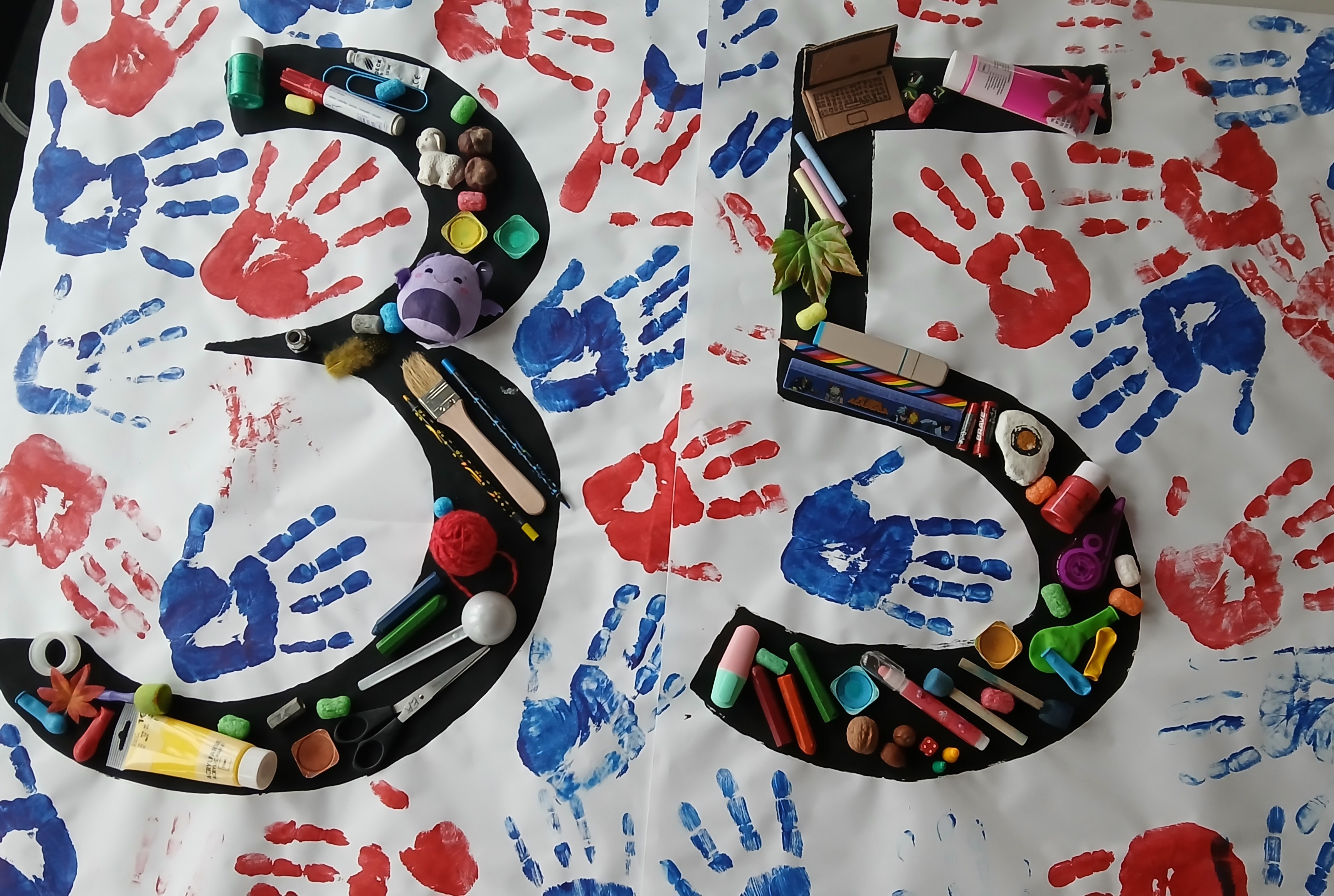 Große „35“ aus schwarzen Pappformen, gefüllt mit Bastelmaterial (Stifte, Pinsel, Knete, kleine Gegenstände) auf weißem Papier, bedeckt mit roten und blauen Handabdrücken.