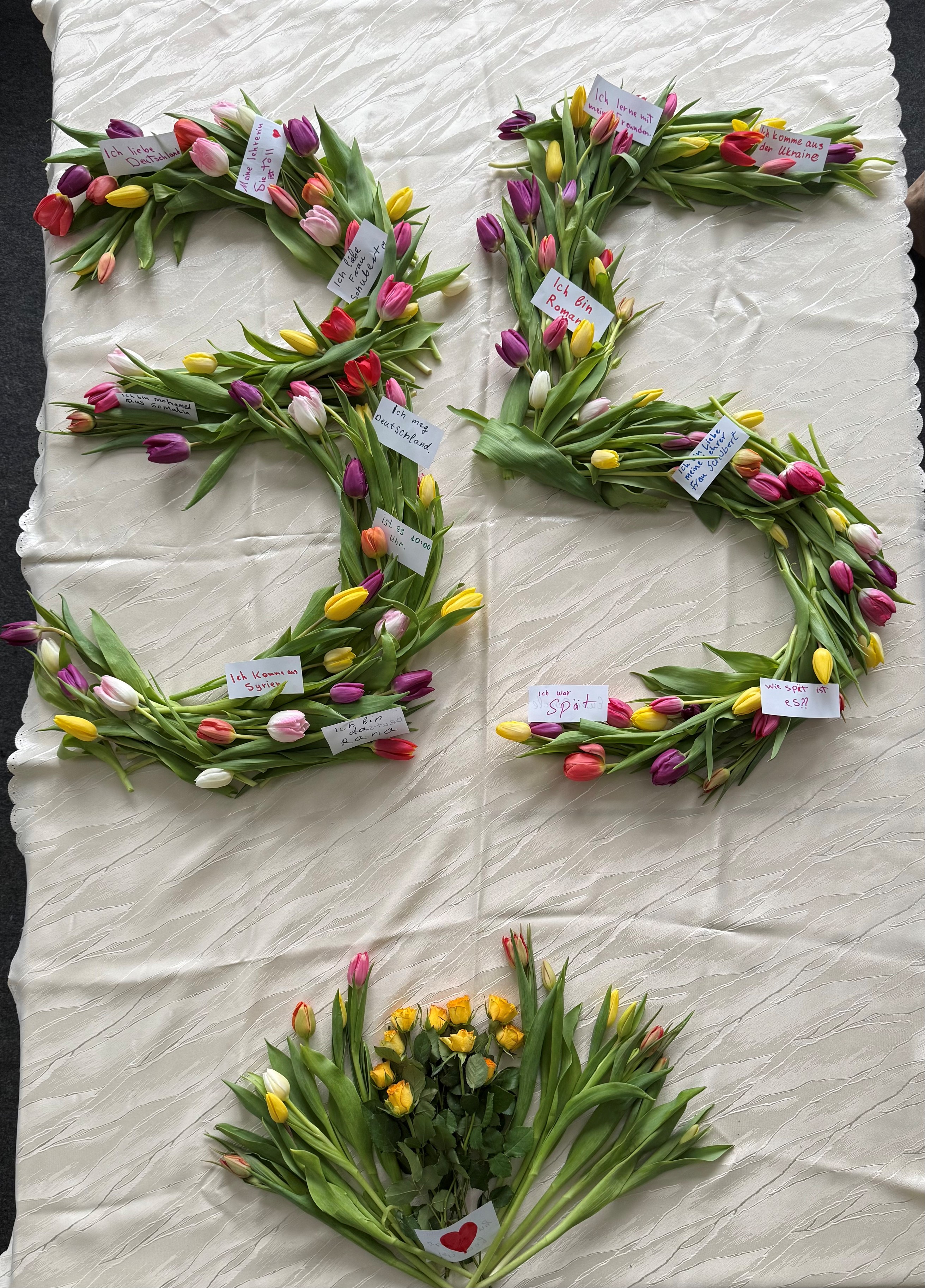 Mit bunten Tulpen und gelben Rosen gelegtes Blumenbild in Form von „B1“ auf einer hellen Stofffläche, ergänzt durch kleine handgeschriebene Zettel mit deutschen Sätzen.