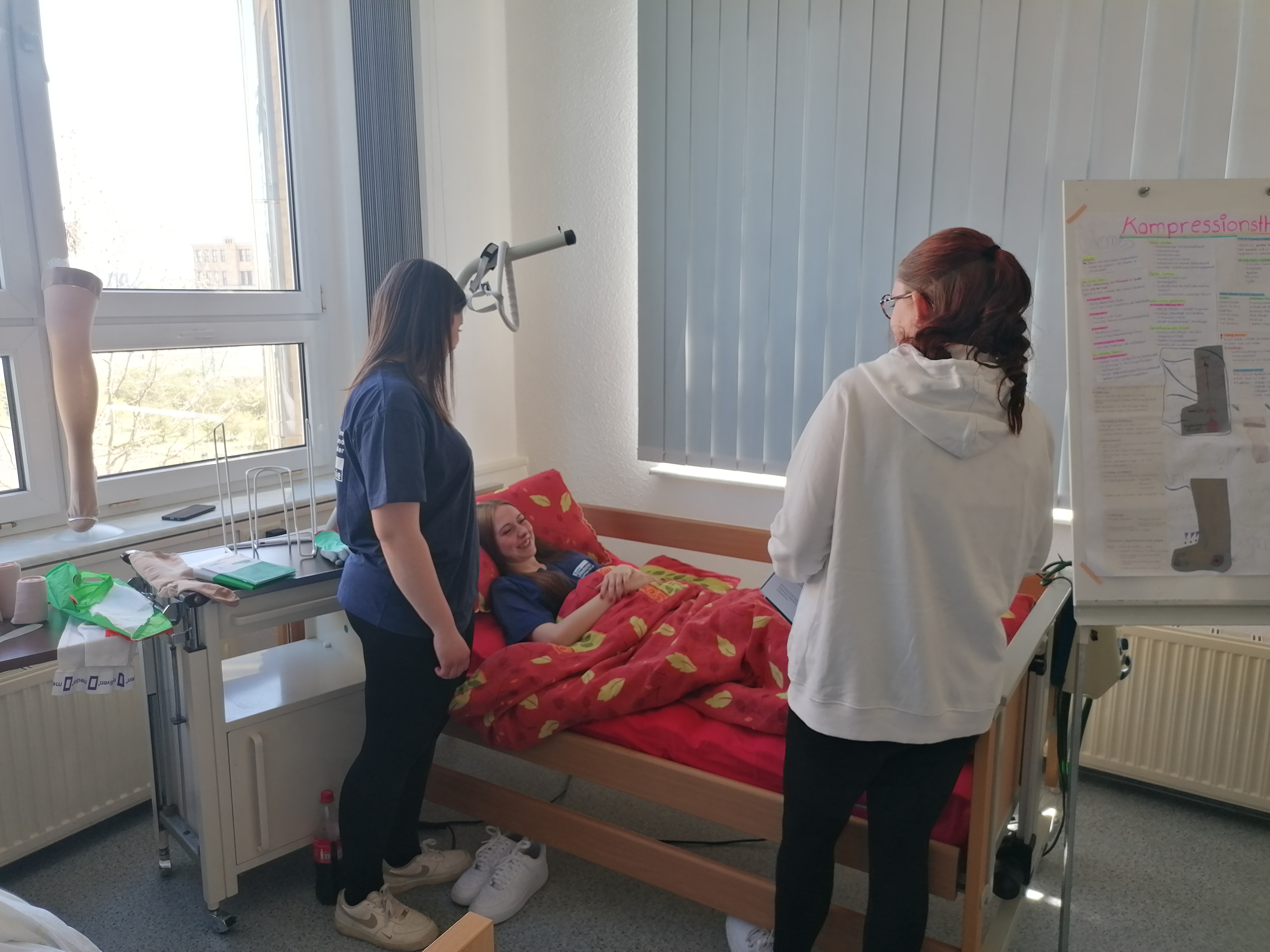 In einem Klassenzimmer liegt eine junge Frau in einem Pflegebett, zwei junge Frauen stehen daneben; rechts ist ein Poster zum Thema "Kompressionsstrümpfe" zu sehen.