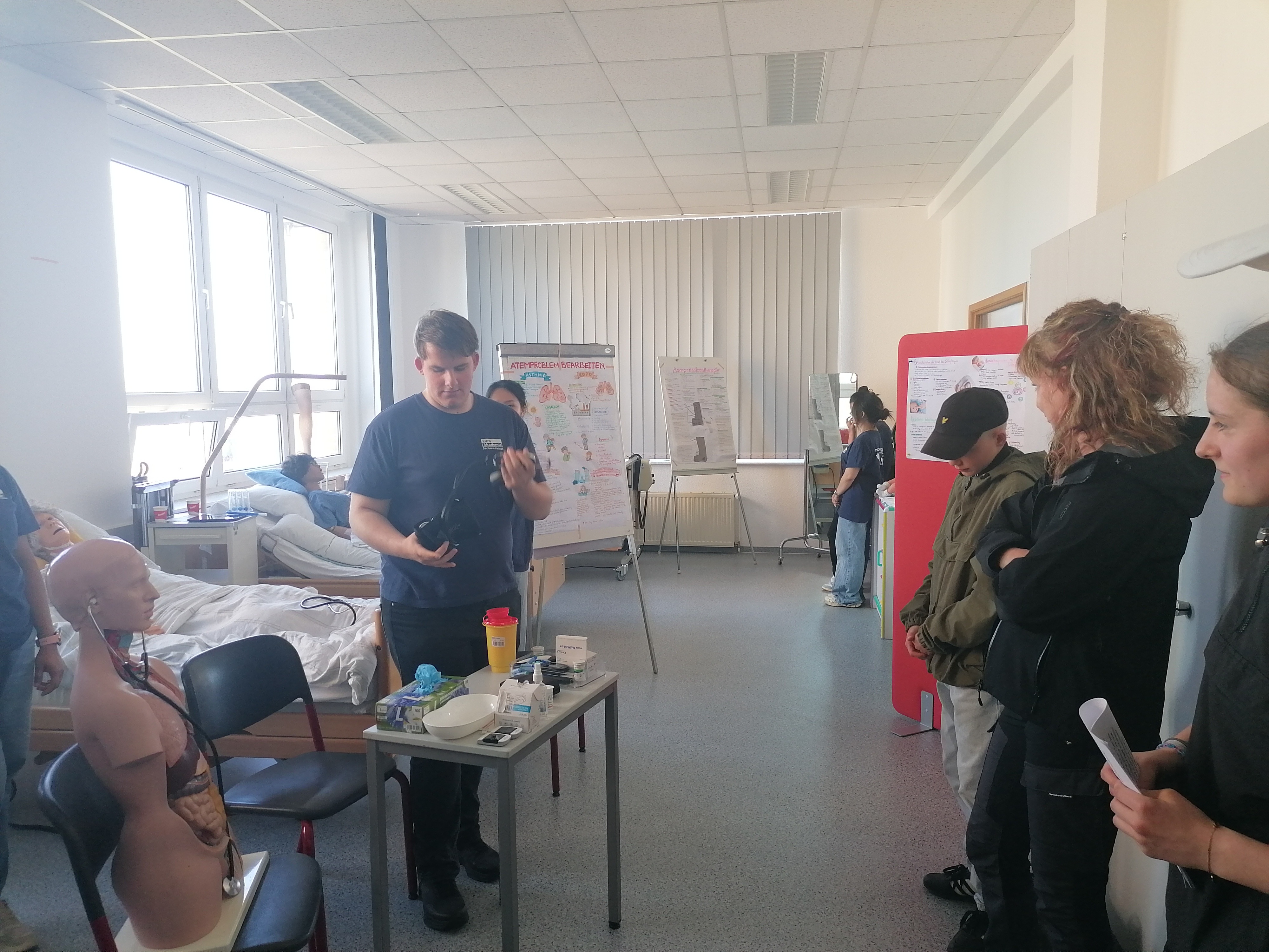 Ein heller Klassenraum: links zwei Pflegebetten mit Puppen; vorne ein junger Mann, der an einem Tisch mit Utensilien etwas demonstriert, während ihm mehrere Menschen zusehen. Im Hintergrund weitere Menschen und Infoplakate.