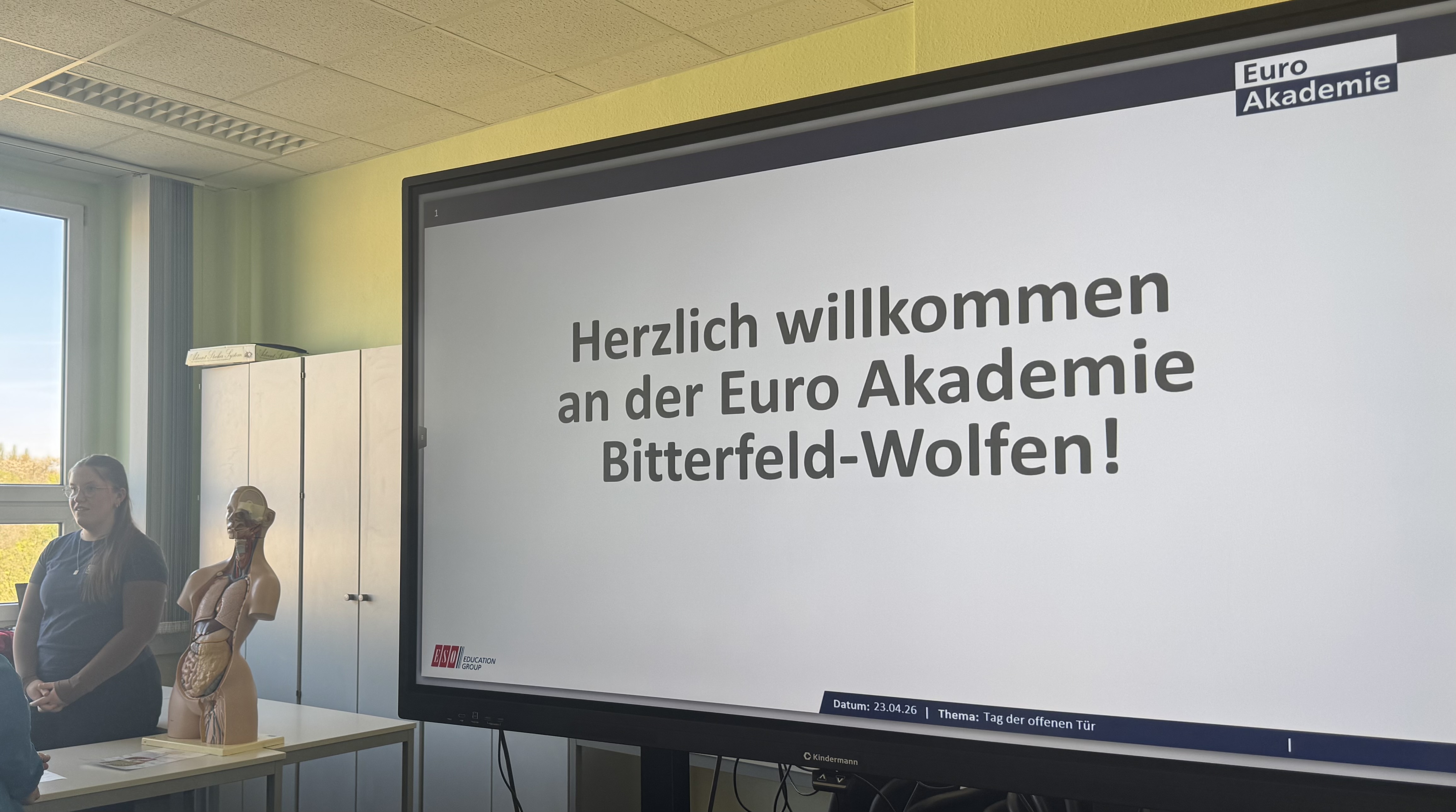 Auf einer weißen Leinwand steht "Herzlich willkommen an der Euro Akademie Bitterfeld-Wolfen!"; links daneben steht eine junge Frau neben dem Modell eines menschlichen Torsos.