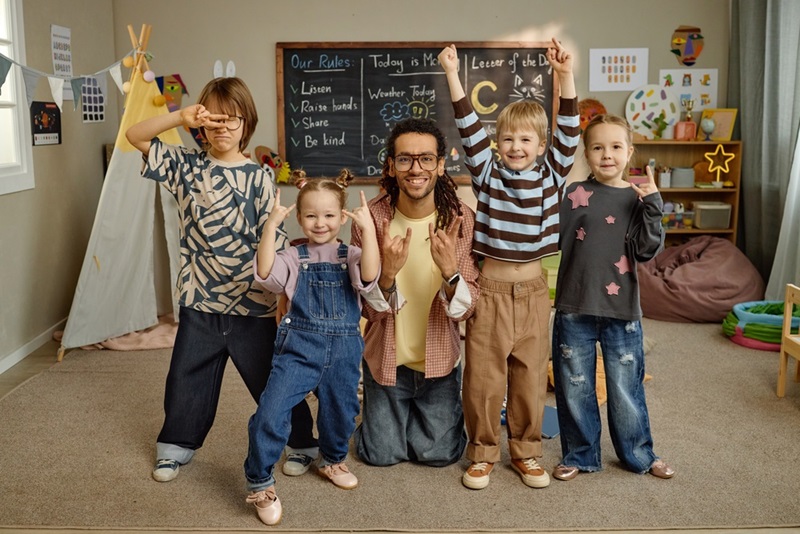 Ein Erwachsener mit Brille und langen, geflochtenen Haaren kniet in einem freundlich eingerichteten Raum zwischen vier Kindern. Alle lächeln und zeigen mit den Händen verschiedene Gesten, die Freude ausdrücken. Im Hintergrund steht eine Tafel mit handgeschriebenen Regeln und Tagespunkten, daneben sind bunte Dekorationen und Spielzeug zu sehen. Die Kinder tragen lässige, farbenfrohe Kleidung. Die Atmosphäre wirkt entspannt und einladend.