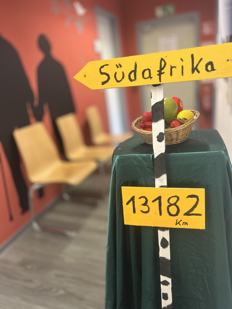 Dekorative Projektstation zum Thema Südafrika im Unterricht der Heilerziehungspflege an der Euro Akademie Chemnitz: Im Vordergrund steht ein gelber Wegweiser mit der Aufschrift ‚Südafrika‘ und dem Hinweis ‚13.182 km‘. Darunter befindet sich ein Korb mit bunt arrangierten künstlichen Früchten, der auf einem mit dunklem Stoff bedeckten Tisch platziert ist. Im Hintergrund sind eine rote Wand, Holzstühle und Silhouetten zweier Menschen zu sehen. Die Szene stellt eine kreative Lernstation dar, die das Thema ‚Reisen mit Klienten‘ visuell und niederschwellig erlebbar macht.