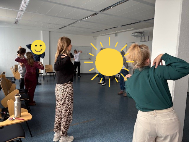 Die Mitarbeitenden stehen in einem großen Raum und führen, im Rahmen des Gesundheitstages der AOK, Übungen durch. Sie tippen sich mit den Händen stehend auf die Schultern.