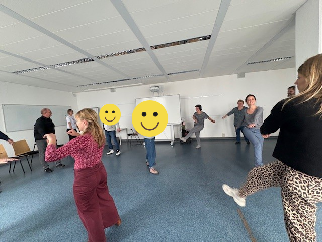 In einem großen Raum stehen die Teilnehmenden des Gesundheitstages im Kreis und heben das gestreckte Bein quer nach oben.