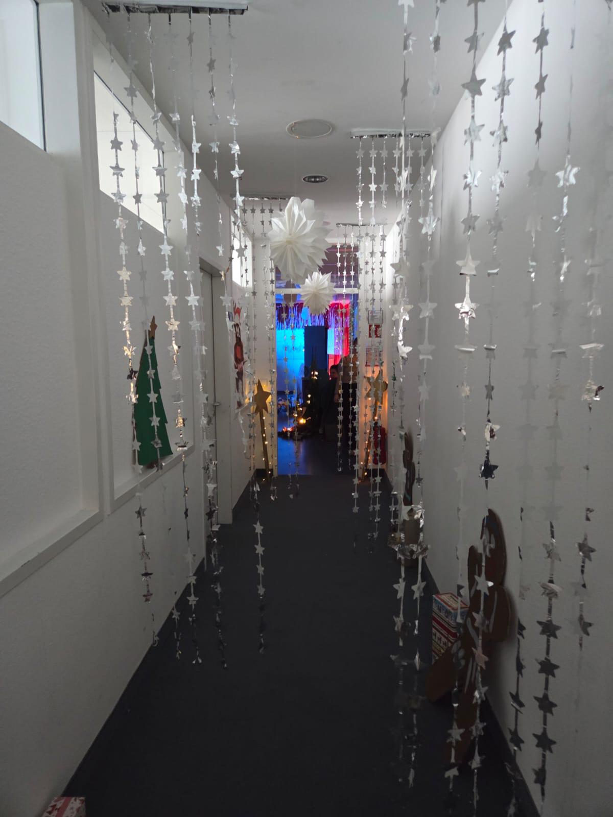 Schmaler, weißer Flur, der mit herunterhängenden silbernen Stern-Girlanden und großen weißen Papiersternen dekoriert ist; am Ende leuchtet ein bunt geschmückter Raum im Hintergrund.