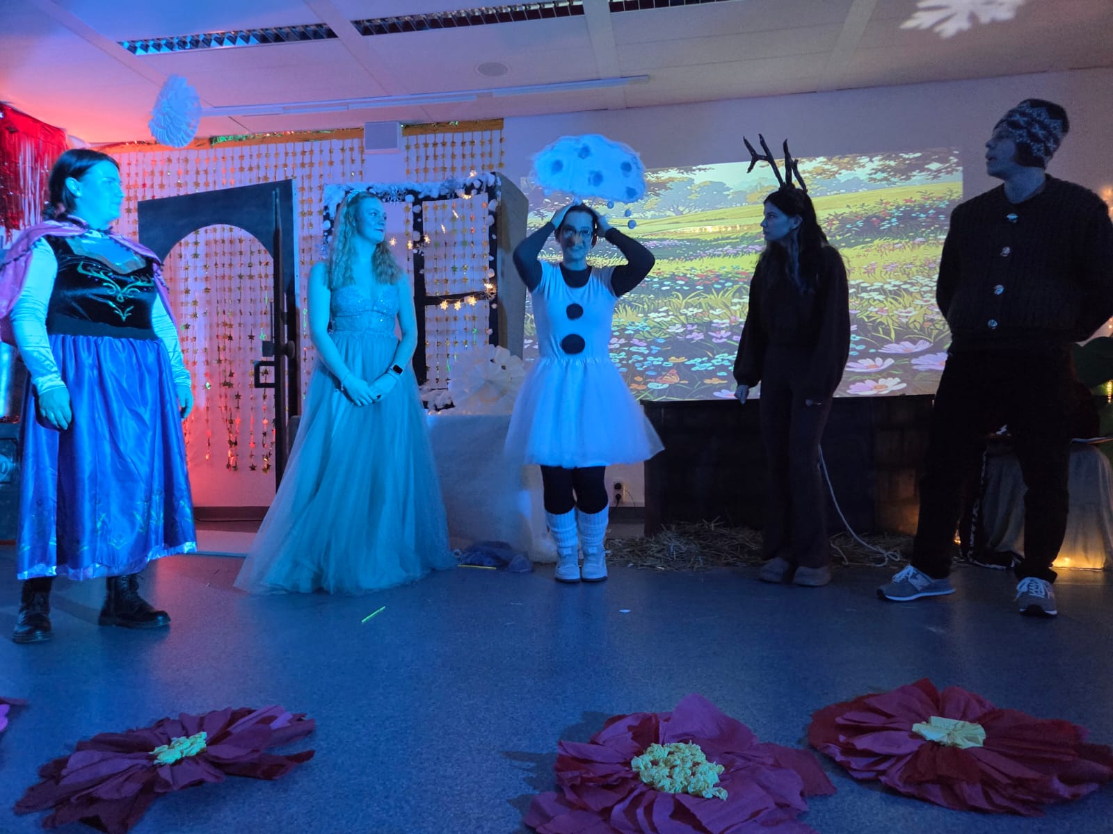 Fünf verkleidete Personen stehen auf einer Bühne in winterlicher „Die Eiskönigin“-Kulisse, mit blauer und roter Beleuchtung, großen Papierflocken und einem projizierten Blumenwiesen-Hintergrund.