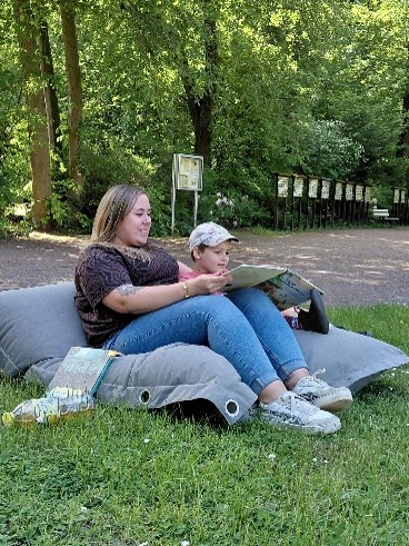 Eine Auszubildende und ein junger Tierparkbesucher sitzen auf einem grauen Sitzkissen im Grünen. Sie halten ein großes Buch auf den Knien aus dem die Auszubildende vorliest. Im Hintergrund stehen Bäume und mehrere Schilder. Die Szene wirkt entspannt und naturverbunden.