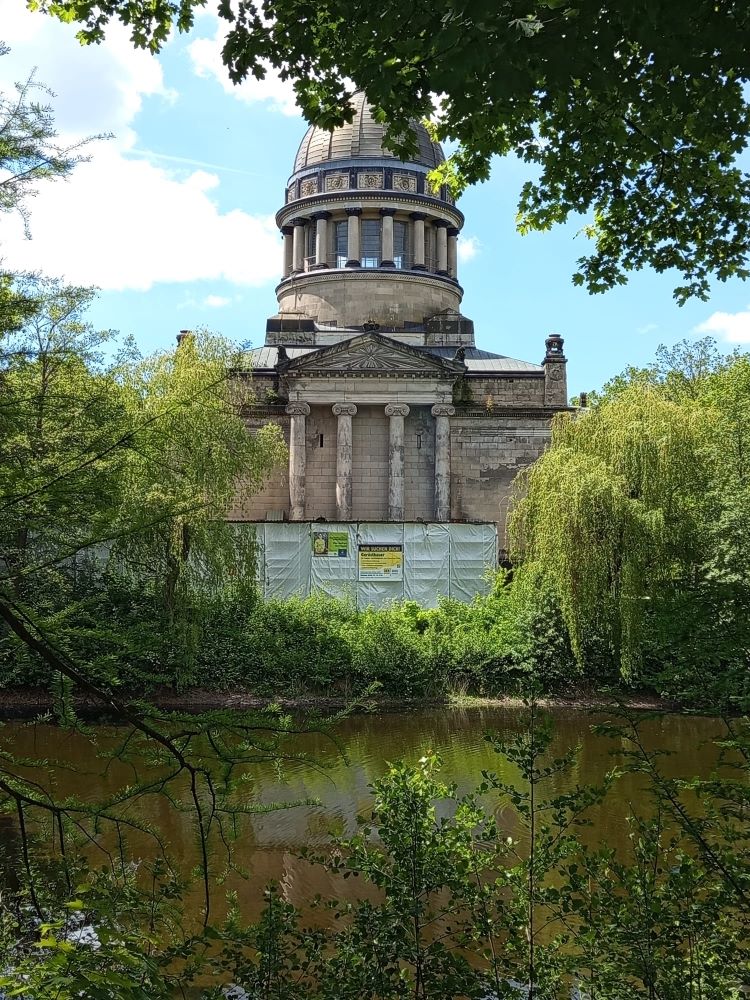 Ein historisches Gebäude mit klassizistischer Fassade und einer großen Kuppel, umgeben von üppigem Grün. Im Vordergrund spiegelt sich das Gebäude in einem ruhigen Teich wider. Das Gebäude ist teilweise mit Gerüsten und Planen abgedeckt, was auf Renovierungsarbeiten hindeutet.