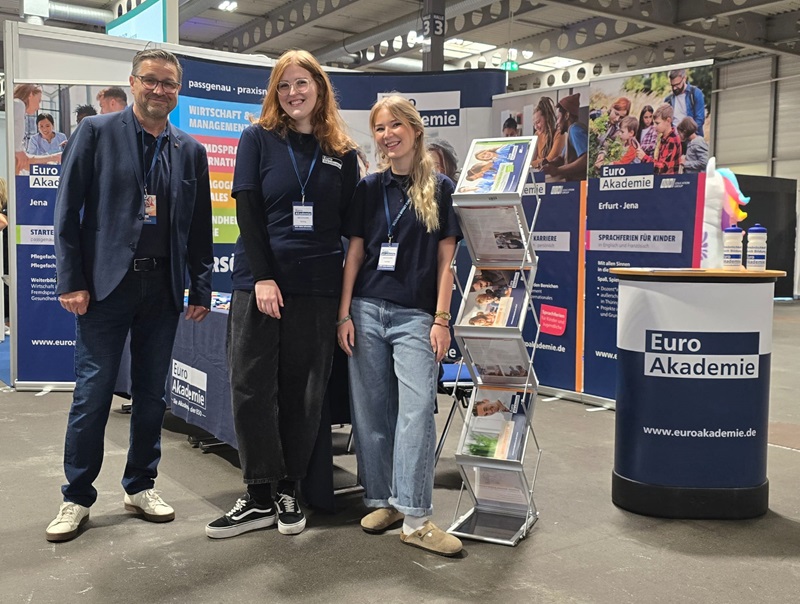 Im Vordergrund stehen drei Personen vor einem Messestand. Sie tragen Namensschilder und dunkelblaue Oberteile. Der Stand ist mit mehreren Roll-up-Bannern, einem Prospektständer mit Broschüren und einem runden Infotresen ausgestattet. Auf dem Tresen und den Bannern ist der Schriftzug 'Euro Akademie' zu sehen. Im Hintergrund sind weitere Messestände und eine große Halle mit sichtbarer Decke und Beleuchtung.