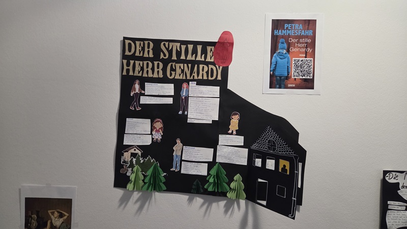 An einer weißen Wand hängt ein großes, schwarzes Plakat mit dem Titel 'DER STILLE HERR GENARDY' in goldenen Buchstaben. Das Plakat zeigt mehrere gezeichnete Figuren, Sprechblasen mit handgeschriebenem Text und ausgeschnittene Bäume. Rechts daneben befindet sich ein weiteres Plakat mit einem blauen Hintergrund, einem Porträtfoto und einem QR-Code. Darauf steht der Name 'PETRA HAMMESFAHR' und der Titel 'Der stille Herr Genardy'.