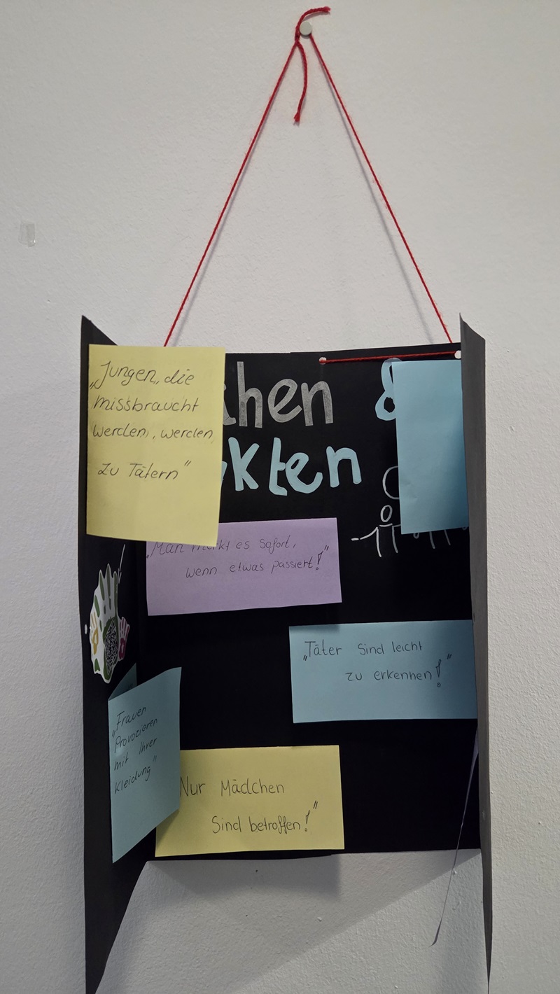 Ein schwarzes Plakat ist an einer roten Schnur aufgehängt. Mehrere bunte Zettel sind darauf befestigt, jeweils mit handgeschriebenem Text. Die Zettel sind in Gelb, Rosa, Blau und Hellblau gehalten. Links unten ist eine kleine, bunte Zeichnung zu sehen. Das Plakat hängt leicht geöffnet von der Wand ab.