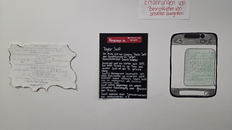 Drei handgefertigte Plakate hängen nebeneinander an einer weißen Wand. Links befindet sich ein unregelmäßig gerissenes Blatt mit handschriftlichem Text. In der Mitte ist ein schwarzes Rechteck mit weißer und roter Schrift, das wie ein Infokasten gestaltet ist. Rechts ist eine gezeichnete Darstellung eines Smartphones mit einem längeren handschriftlichen Text auf dem Display. Oberhalb der Plakate hängt ein weiteres Blatt mit roter Handschrift.