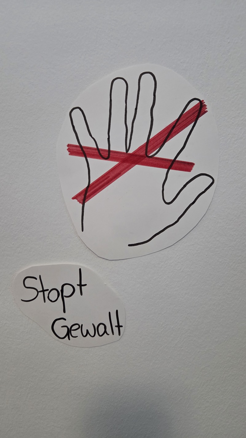 Eine stilisierte Hand ist mit schwarzer Linie auf weißem Papier gezeichnet. Zwei dicke, rote Linien kreuzen sich in Form eines X über der Handfläche. Darunter steht auf einem separaten weißen Papierstück in klarer Handschrift: 'Stopt Gewalt'. Der Hintergrund ist einfarbig und hell.