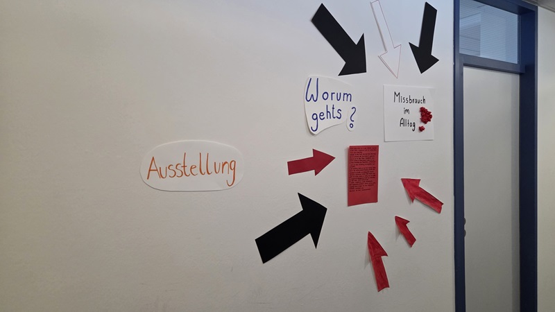 Weiße Wand mit mehreren Pfeilen aus schwarzem und rotem Papier, die auf verschiedene Bereiche zeigen. Links steht das Wort 'Ausstellung' in orange auf einem weißen Papier. Rechts oben befindet sich ein Schild mit der handschriftlichen Frage 'Worum gehts?' und daneben ein weiteres Schild mit der Aufschrift 'Missbrauch im Alltag', das mit roten Papierblumen verziert ist. In der Mitte ist ein rotes Blatt mit handgeschriebenem Text, das von mehreren roten Pfeilen umgeben ist. Rechts ist ein blauer Türrahmen sichtbar.