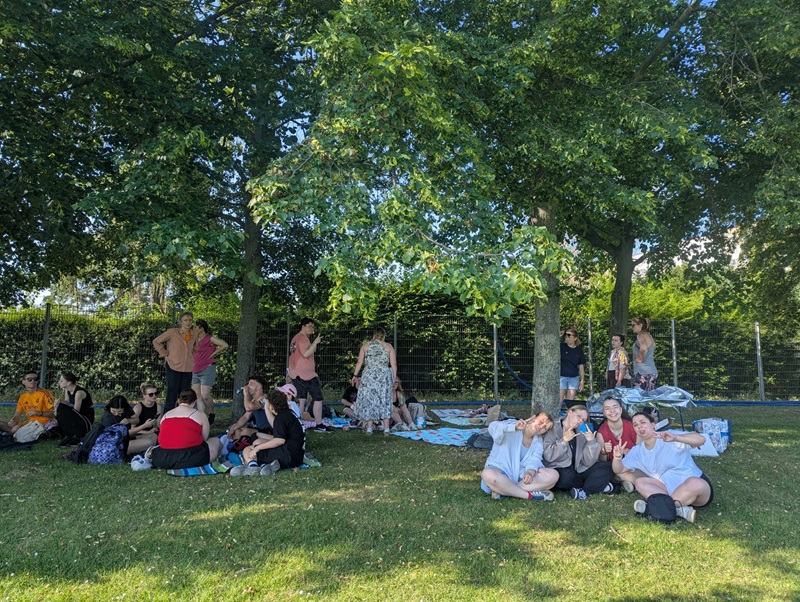 Die Azubis sitzen und stehen auf einer Wiese unter großen, schattenspendenden Bäumen. Einige Personen sitzen auf Decken, andere lehnen an Baumstämmen oder unterhalten sich im Stehen. Im Vordergrund sitzt eine Gruppe junger Menschen eng beieinander und lächelt in die Kamera. Im Hintergrund verläuft ein Zaun entlang einer dichten Hecke. Die Szene wirkt entspannt und sommerlich.