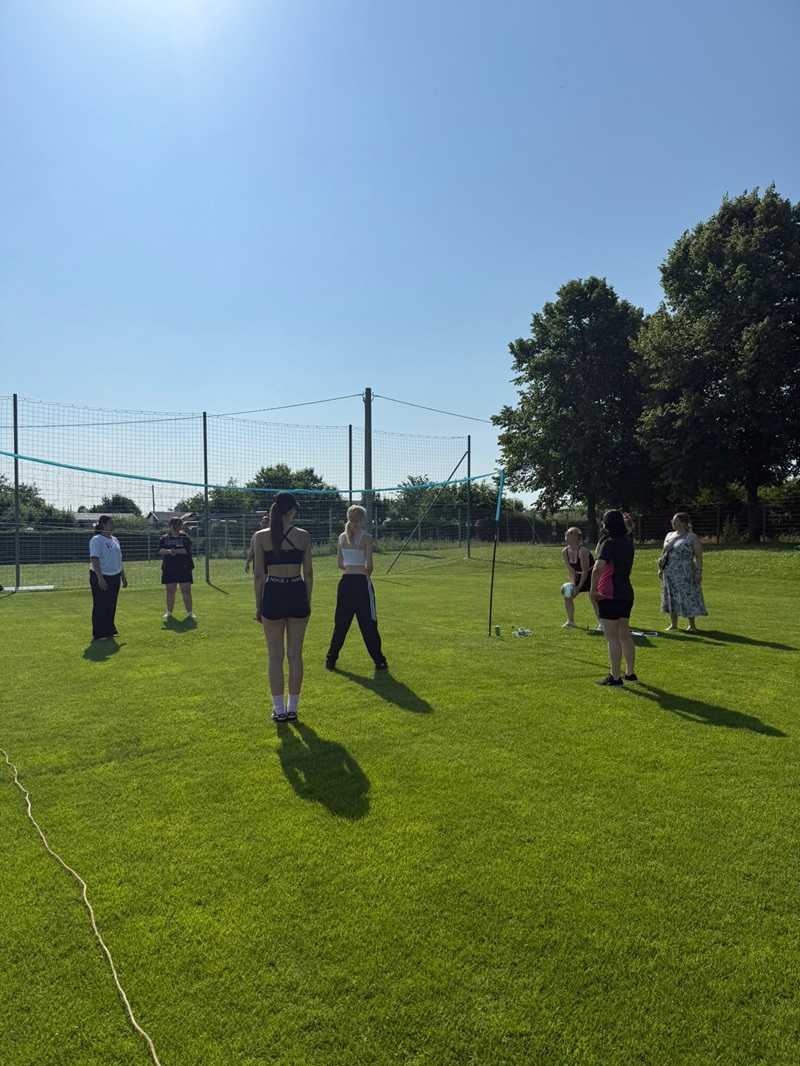 Mehrere Auszubildende stehen auf einem gepflegten Rasenplatz vor einem Volleyballnetz. Sie tragen sportliche Sommerkleidung und stehen locker verteilt, als würden sie sich auf ein Spiel vorbereiten oder unterhalten. Im Hintergrund sind Bäume und ein Zaun zu sehen, der den Platz begrenzt. Die Sonne scheint, der Himmel ist wolkenlos und hellblau.