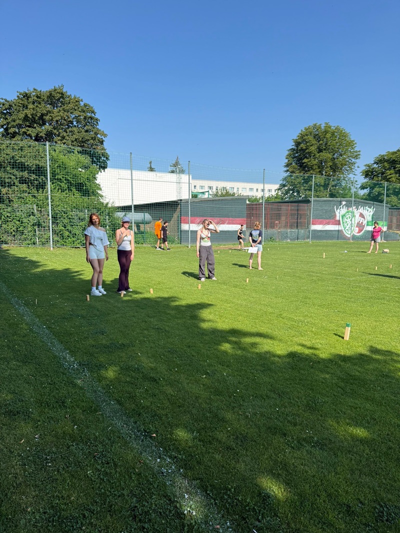 Auf dem Sportplatz stehen die Azubis, im Hintergrund sind Bäume, ein Gebäude mit weißer Fassade und ein rot-weißes Wandbild zu sehen. Die Personen tragen sportliche Freizeitkleidung und verteilen sich locker auf dem Rasen. Auf dem Boden sind kleine Holzklötze aufgestellt, die auf ein Wurfspiel hindeuten.