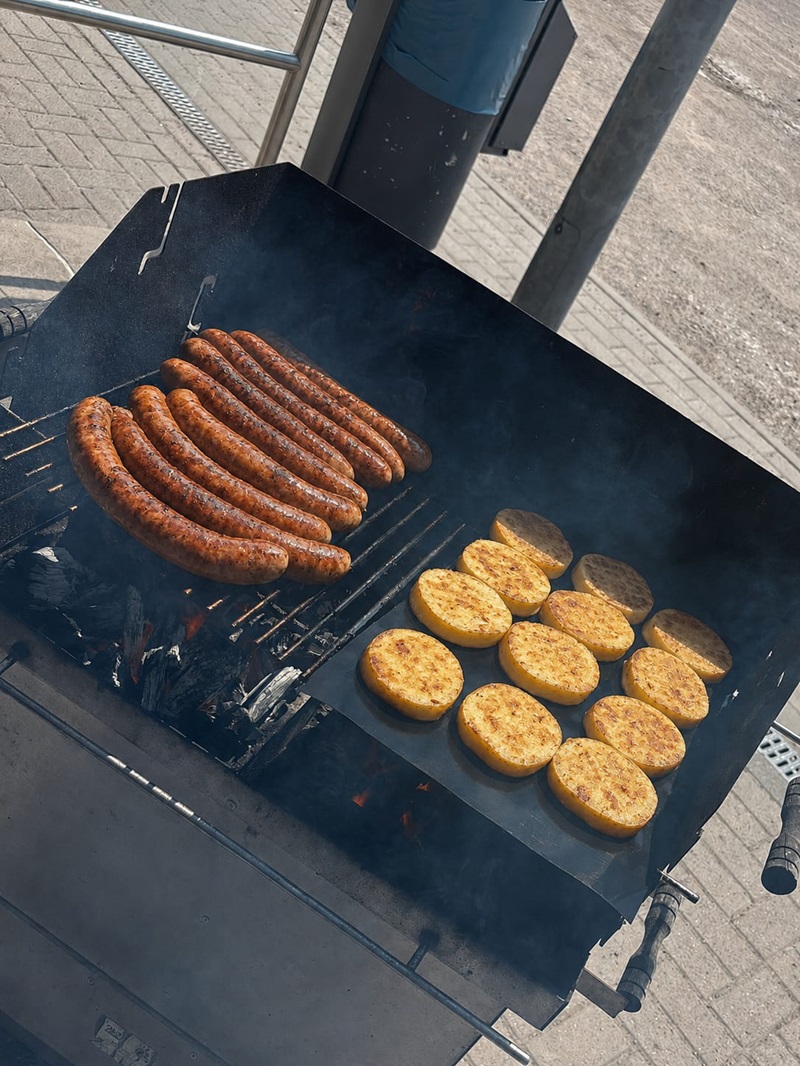 Auf einem Grill liegen mehrere Bratwürste nebeneinander auf dem Rost, daneben befinden sich runde, goldbraun gebratene Kartoffelscheiben auf einer Grillplatte. Im Hintergrund sind Pflastersteine und ein Teil eines Metallpfostens zu sehen. Rauch steigt vom Grill auf.