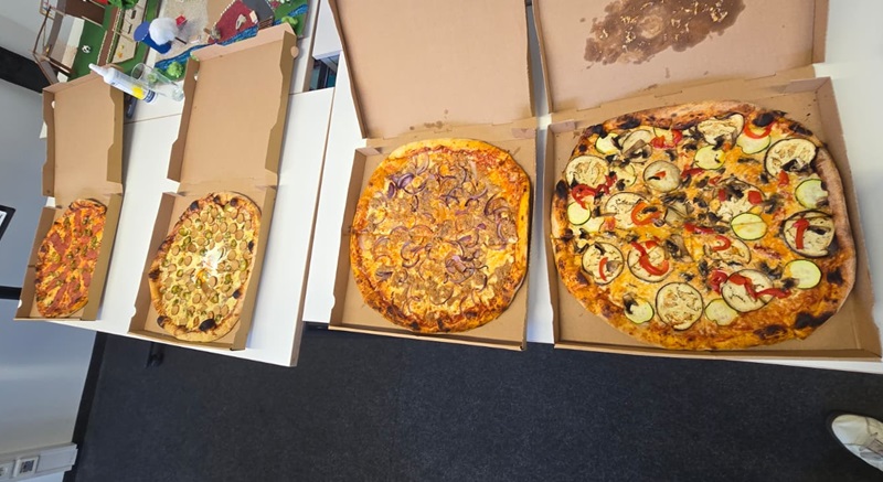 Vier geöffnete Pizzakartons stehen nebeneinander auf einer weißen Tischfläche. Jede Pizza hat einen anderen Belag, darunter verschiedene Gemüsesorten, Pilze, Zwiebeln und Paprika. Die Pizzen sind rund und groß, der Teig ist goldbraun gebacken. Im Hintergrund sind Teile eines Büros mit Stühlen, einem Schreibtisch und einigen Gegenständen zu erkennen.
