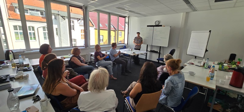In einem hellen Besprechungsraum mit großen Fenstern sitzen die Mitarbeitenden im Halbkreis auf Stühlen. Eine Person steht vorne neben einem Whiteboard und spricht zur Gruppe. Auf den Tischen liegen verschiedene Getränke, Schreibutensilien und Unterlagen. Im Hintergrund sind Flipcharts und eine Uhr an der Wand zu sehen. Durch die Fenster fällt Tageslicht und es sind Dächer benachbarter Gebäude sichtbar.