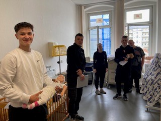 Zu sehen sind vier Schüler und eine Schülerin in einem Klassenraum, die in die Kamera lächeln. Links im Bild steht einer der Schüler mit einer Babypuppe im Arm. Hinter ihm befindet sich ein Kinderbett.