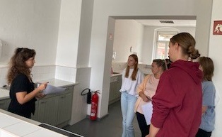 Zu sehen sind sechs Schülerinnen, die auf dem Flur einer Schule im Kreis stehen und miteinander diskutieren. Im Hintergrund ist ein Durchgangsbogen des Flurs zu sehen.