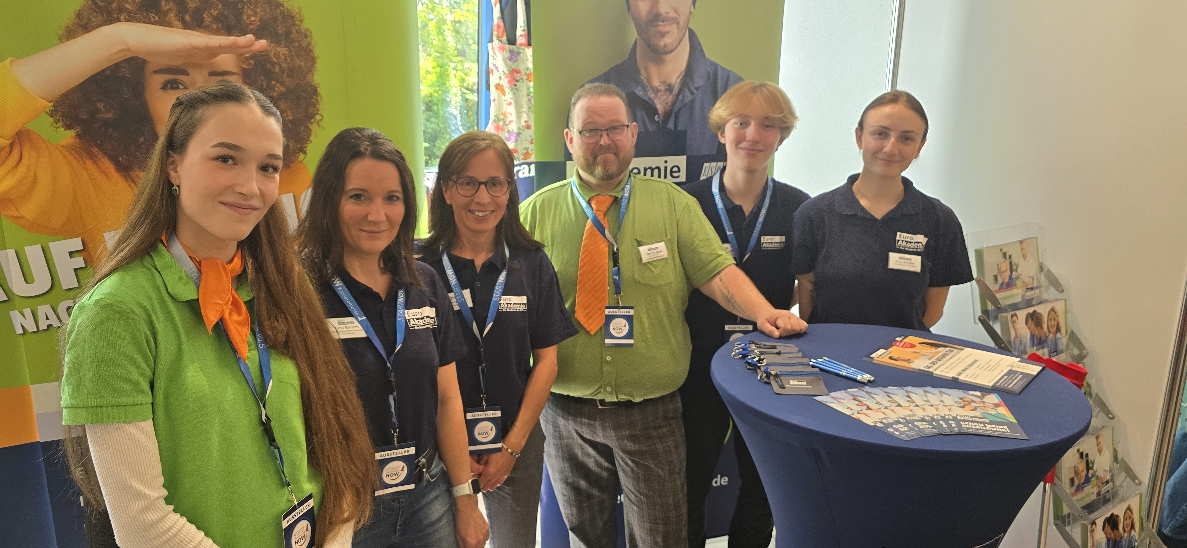 Sechs Mitarbeitende und Auszubildende der Euro Akademie stehen lächelnd an einem Messestand hinter einem blauen Stehtisch mit Flyern, Kugelschreibern und Schlüsselbändern vor farbigen Roll-ups.