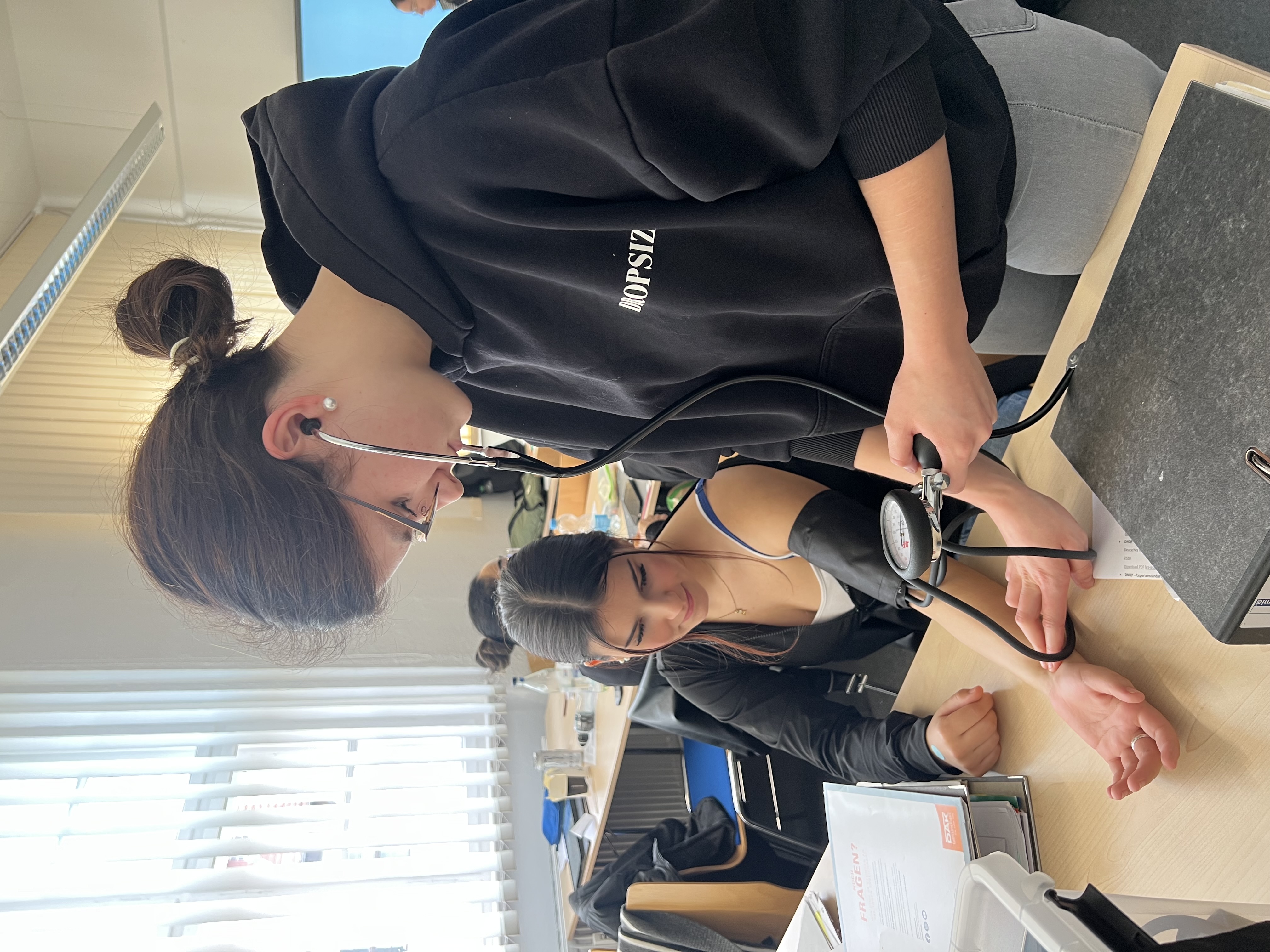 In einem Unterrichtsraum misst eine Schülerin mit Stethoskop und Blutdruckmanschette den Blutdruck einer Mitschülerin, die mit freiem Arm am Tisch sitzt.