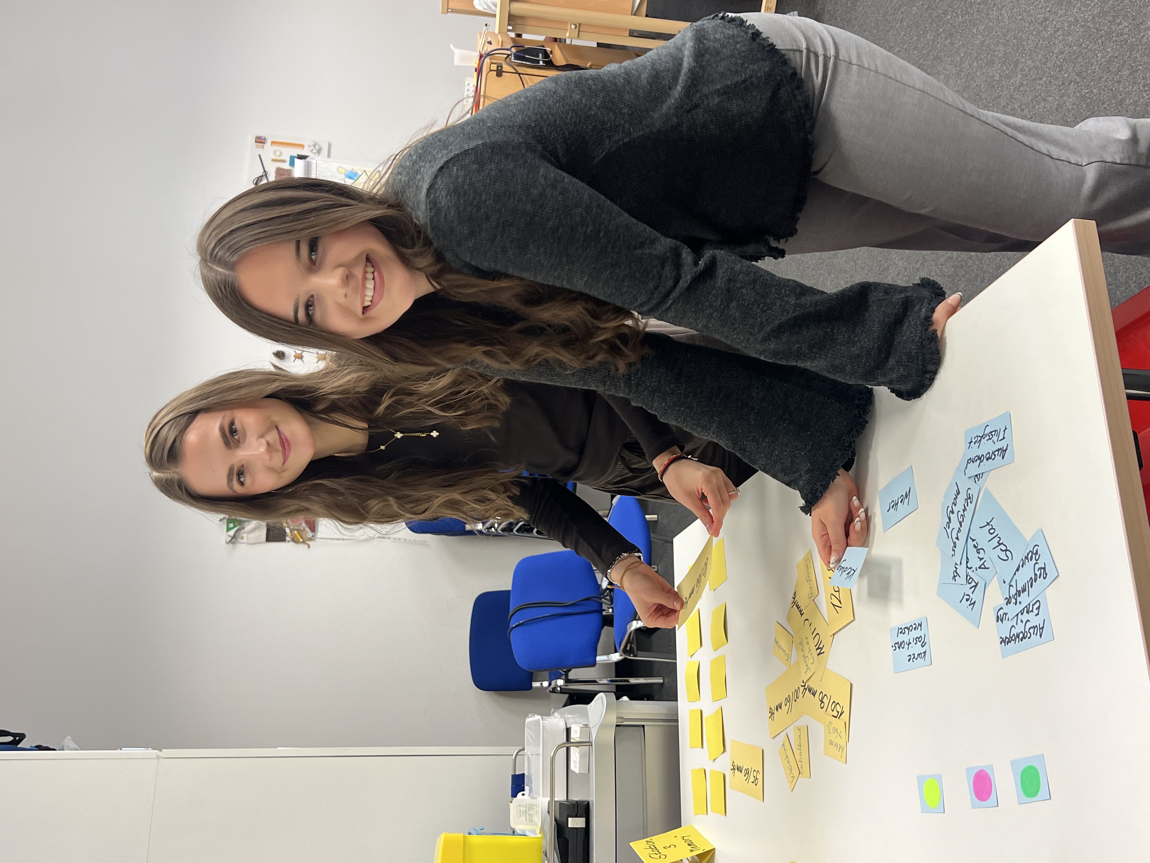 Zwei junge Frauen stehen lächelnd an einem Tisch und ordnen bunte Haftnotizen mit Stichworten für eine Gruppenarbeit.