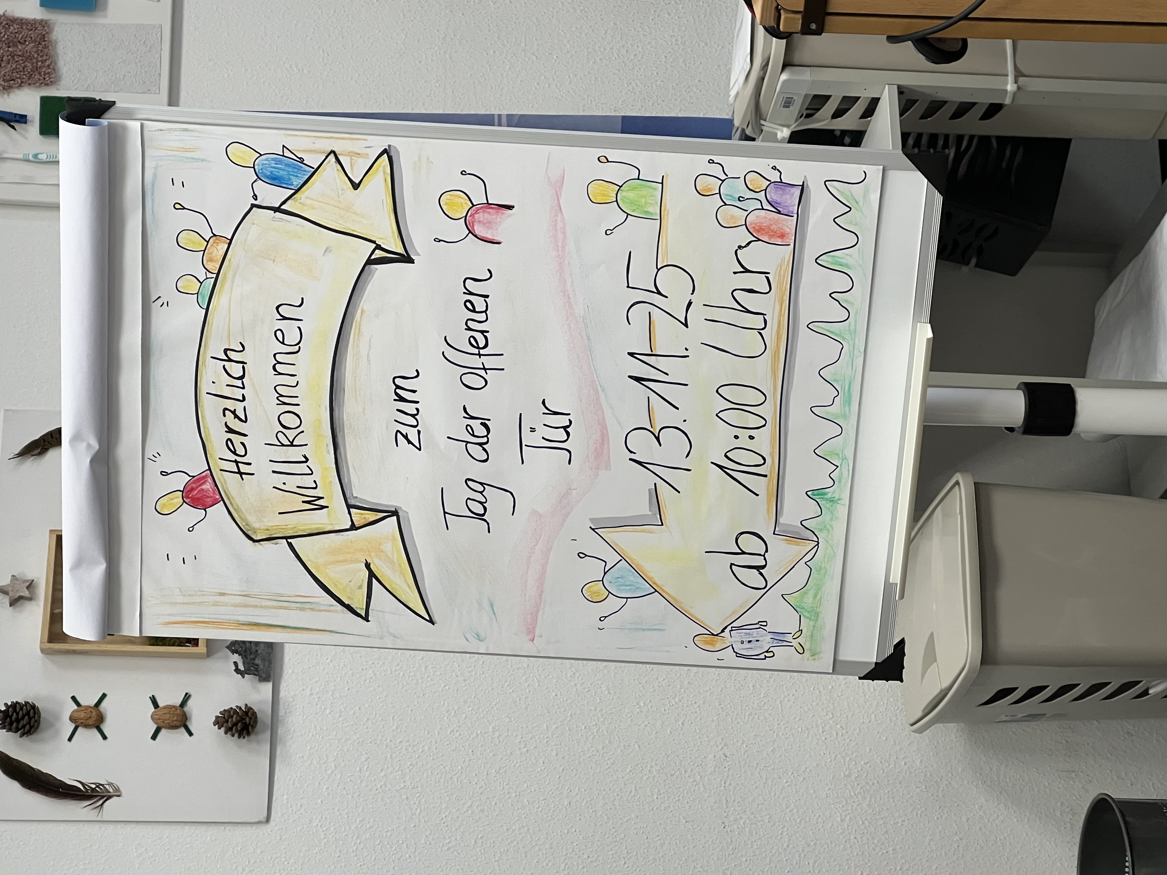 Handgezeichnetes Flipchart mit der Aufschrift „Herzlich Willkommen zum Tag der offenen Tür 13.11.25 ab 10:00 Uhr“, verziert mit Figuren und einem großen Pfeil, steht in einem Fachraum.