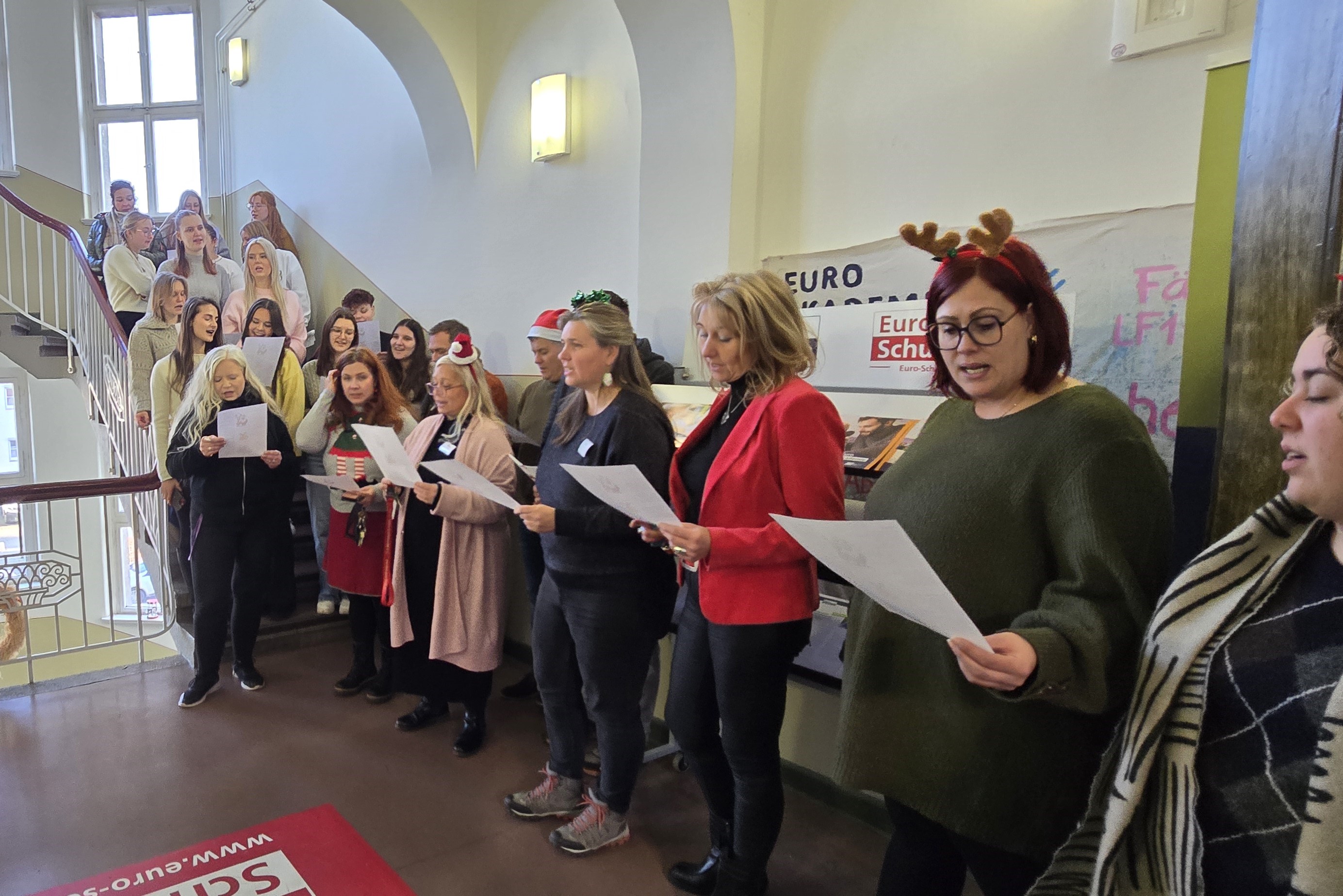 Eine große Gruppe von Lehrkräften und Studierenden steht im Treppenhaus der Euro Akademie, viele mit Weihnachtsaccessoires, und singt gemeinsam mit Notenblättern in der Hand.