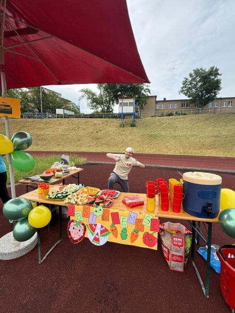 Auf einem Sportplatz steht ein bunt verzierter Stand mit Obst, Gemüse und Getränken. Dahinter steht ein junger Mann, der beide Daumen in die Höhe hält.