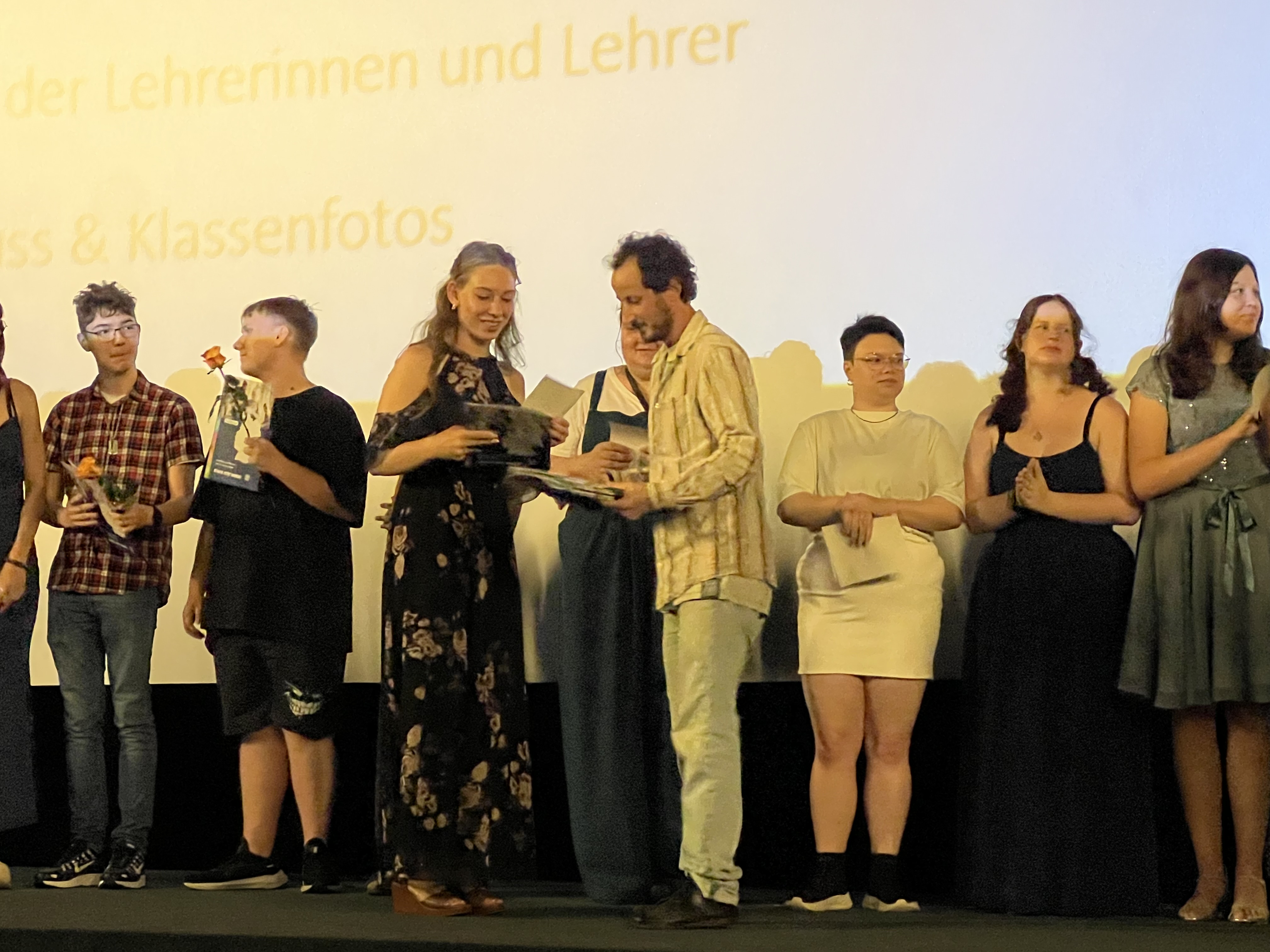 Auf einer Bühne steht eine Reihe junger Menschen, die gerade ihre Zeugnisse überreicht bekommen.