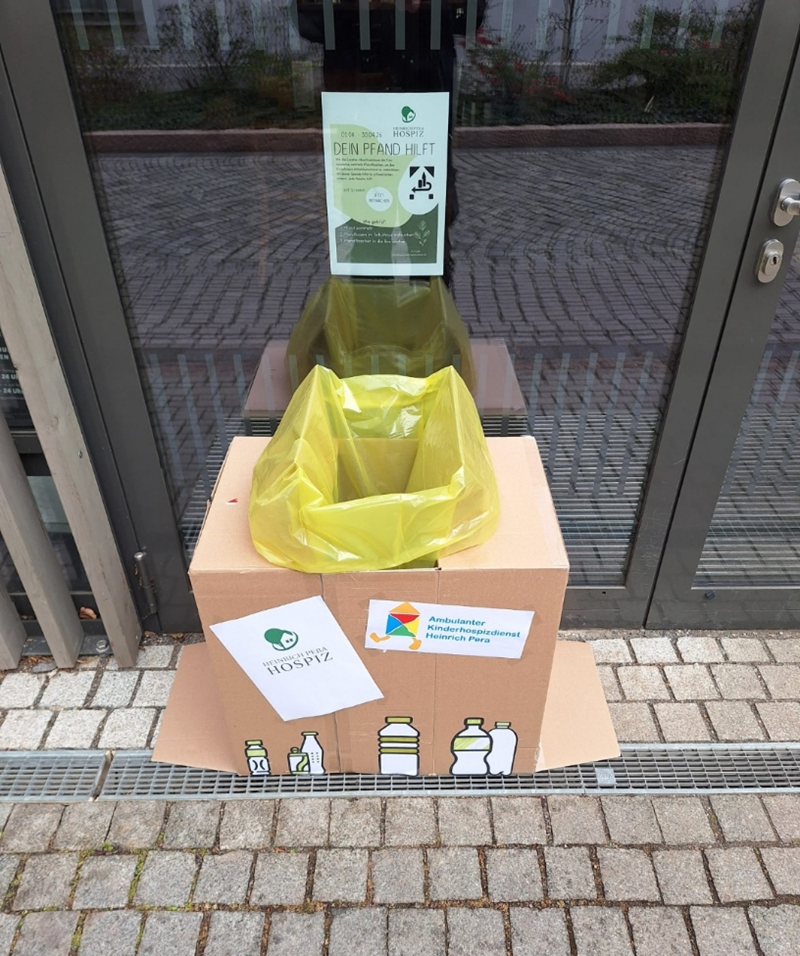 Ein großer, rechteckiger Karton steht auf einem gepflasterten Boden vor einer Glastür. Oben ist ein gelber Müllbeutel in die Öffnung des Kartons eingespannt. Auf der Vorderseite sind stilisierte Flaschen und Behälter in verschiedenen Formen abgebildet. Zwei Blätter sind an der Vorderseite befestigt: Eines zeigt ein rundes Logo mit Text, das andere ein farbiges Logo mit Schriftzug. Im Hintergrund hängt ein weiteres Plakat an der Tür.