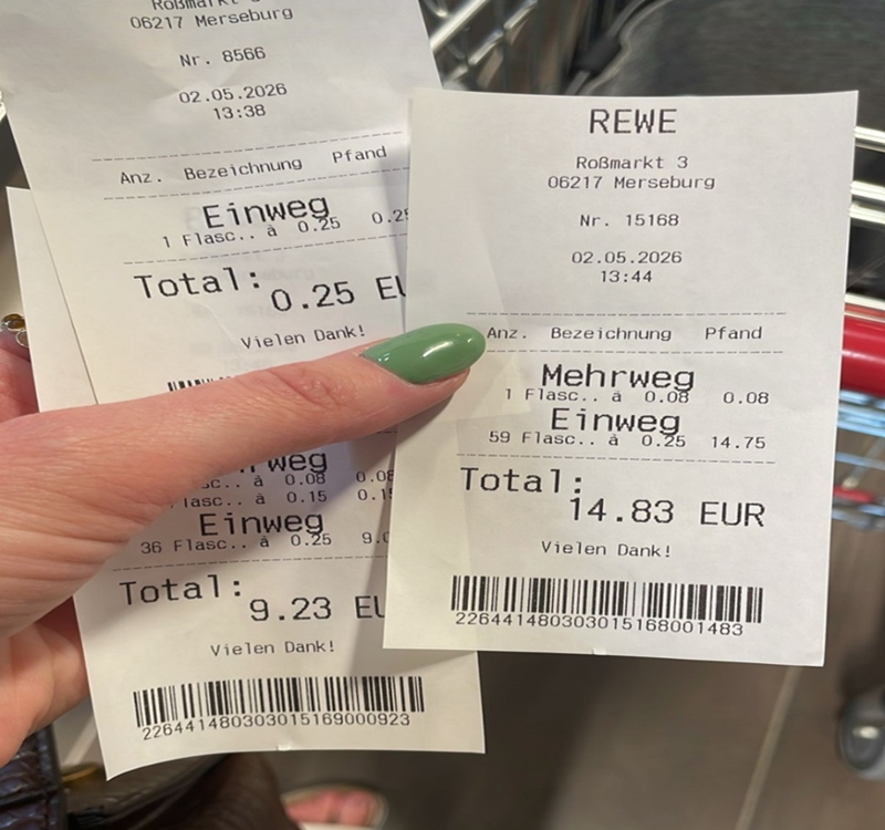 Zwei Kassenbons werden von einer Hand mit grün lackierten Fingernägeln gehalten. Beide Bons stammen von einem Supermarkt und zeigen detaillierte Informationen zu gekauften Mehrweg- und Einwegflaschen, Pfandbeträgen, Gesamtpreisen und dem Einkaufsdatum. Im Hintergrund ist ein Einkaufswagen zu erkennen.