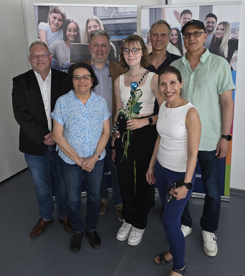 Die Absolventin Lara trägt eine Brille und eine helle Hose hält eine einzelne blaue Rose und steht in der Mitte einer Gruppe von Mitarbeitenden der Euro Akademie Hannover, die gemeinsam mit ihr für ein Erinnerungsfoto posieren. Die Personen tragen überwiegend Freizeitkleidung und lächeln in die Kamera. Im Hintergrund sind zwei Roll-Ups mit großformatigen Fotos von jungen Menschen und dezenten Farbstreifen zu sehen. Die Szene spielt in einem Innenraum mit grauem Boden.