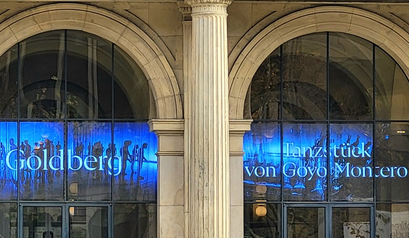 Zwei große, nebeneinanderliegende Bogenfenster des historischen Gebäudes des Opernhauses Hannover mit hellen Sandsteinsäulen dazwischen. Hinter den Fenstern leuchtet ein blaues Licht, das die Silhouetten von Tänzern in verschiedenen Posen zeigt. Auf den Fenstern sind weiße Schriftzüge zu lesen: links 'Goldberg' und rechts 'Tanzstück von Goyo Montero'. Die Szene zeigt die Ankündigung der Ballettaufführung "Goldberg".