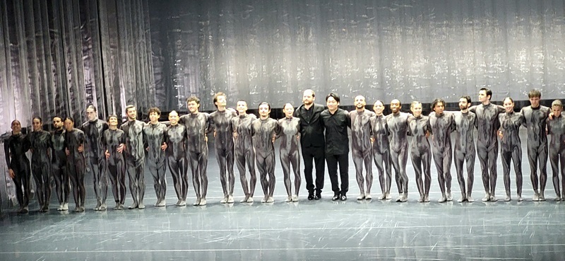 Eine Gruppe von Balletttänzerinnen und -tänzern steht in einer Reihe auf der Bühne des Opernhauses Hannover. Sie tragen eng anliegende, silbergraue Kostüme mit einem metallischen Schimmer, die ihre Körperformen betonen. Die Bühne ist mit einem Vorhang aus silbrig glänzendem Stoff im Hintergrund ausgestattet, der eine elegante Atmosphäre schafft. In der Mitte der Reihe stehen zwei Personen in schwarzer Kleidung, die sich von den Tänzern abheben. Die Tänzerinnen und Tänzer halten sich an den Händen und blicken nach vorne. Die Szene zeigt den Schlussapplaus des Balletts „Goldberg“, das von Schülerinnen und Schülern der Euro Akademie Hannover besucht wurde.