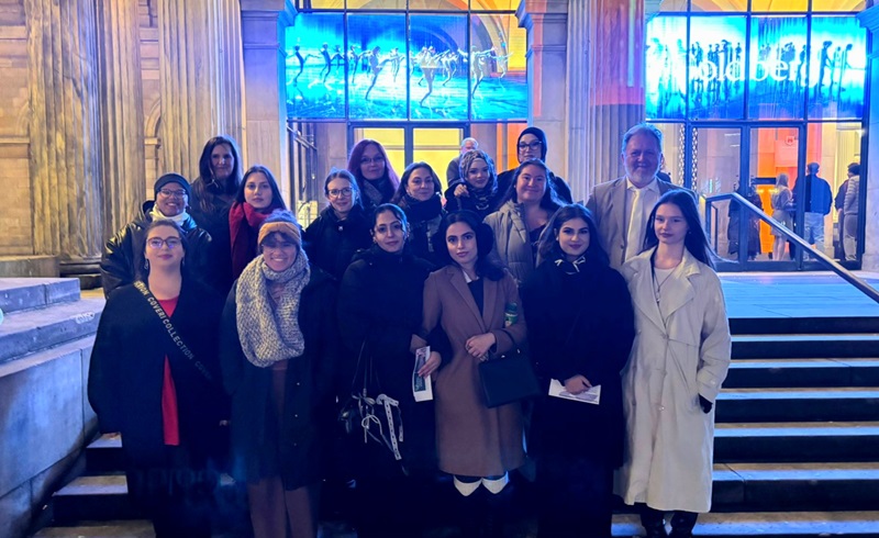 Die Schülerinnen und Schülern der Euro Akademie Hannover stehen vor dem beleuchteten Eingangsbereich auf einer Treppe des Opernhauses Hannover und posieren für ein Gruppenfoto. Sie tragen unterschiedliche Winterbekleidung, darunter Mäntel, Schals und Jacken in verschiedenen Farben wie Beige, Schwarz und Dunkelblau. Im Hintergrund sind große Glasfenster mit blauer Beleuchtung und der Schriftzug "Goldberg" sichtbar, der auf das Ballettstück hinweist, das sie besucht haben.