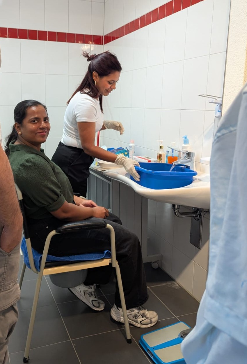 Eine Schülerin sitzt vor einem Waschbecken auf einem Stuhl. Eine zweite Schülerin mit Einweg-Handschuhen trifft Vorbereitungen, um sie zu waschen: Sie lässt Wasser in eine viereckige Plastikschüssel fließen und hat die Pflegeprodukte schon auf dem Tisch bereitgestellt.