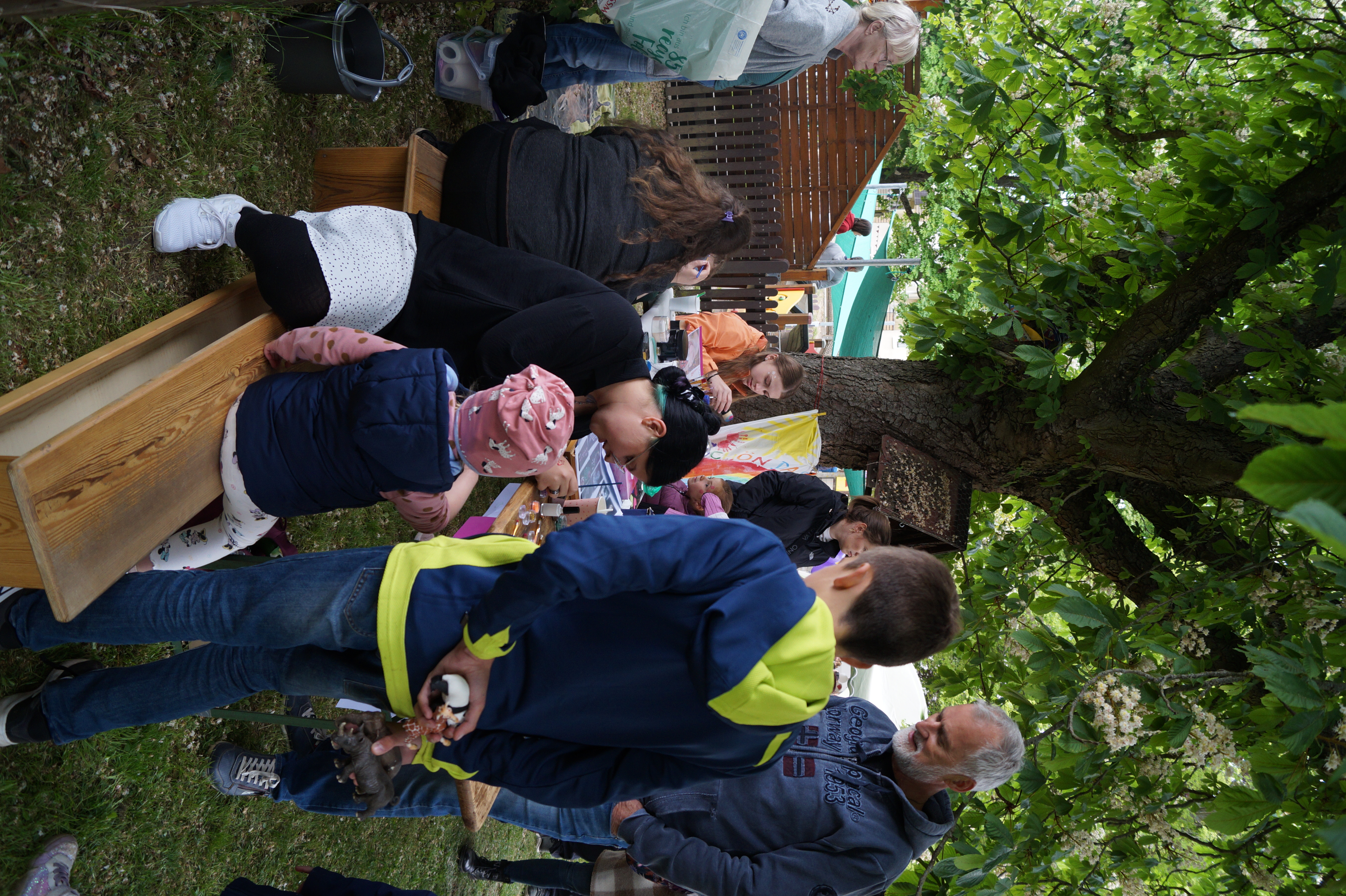 Personen sitzen und stehen um einen Tisch unter einem Baum herum, an dem offenbar Kinderschminken angeboten wird. Erwachsene schminken Kinder, während weitere Personen im Hintergrund stehen. Die Atmosphäre ist entspannt und gesellig auf einer Grünfläche im Freien.