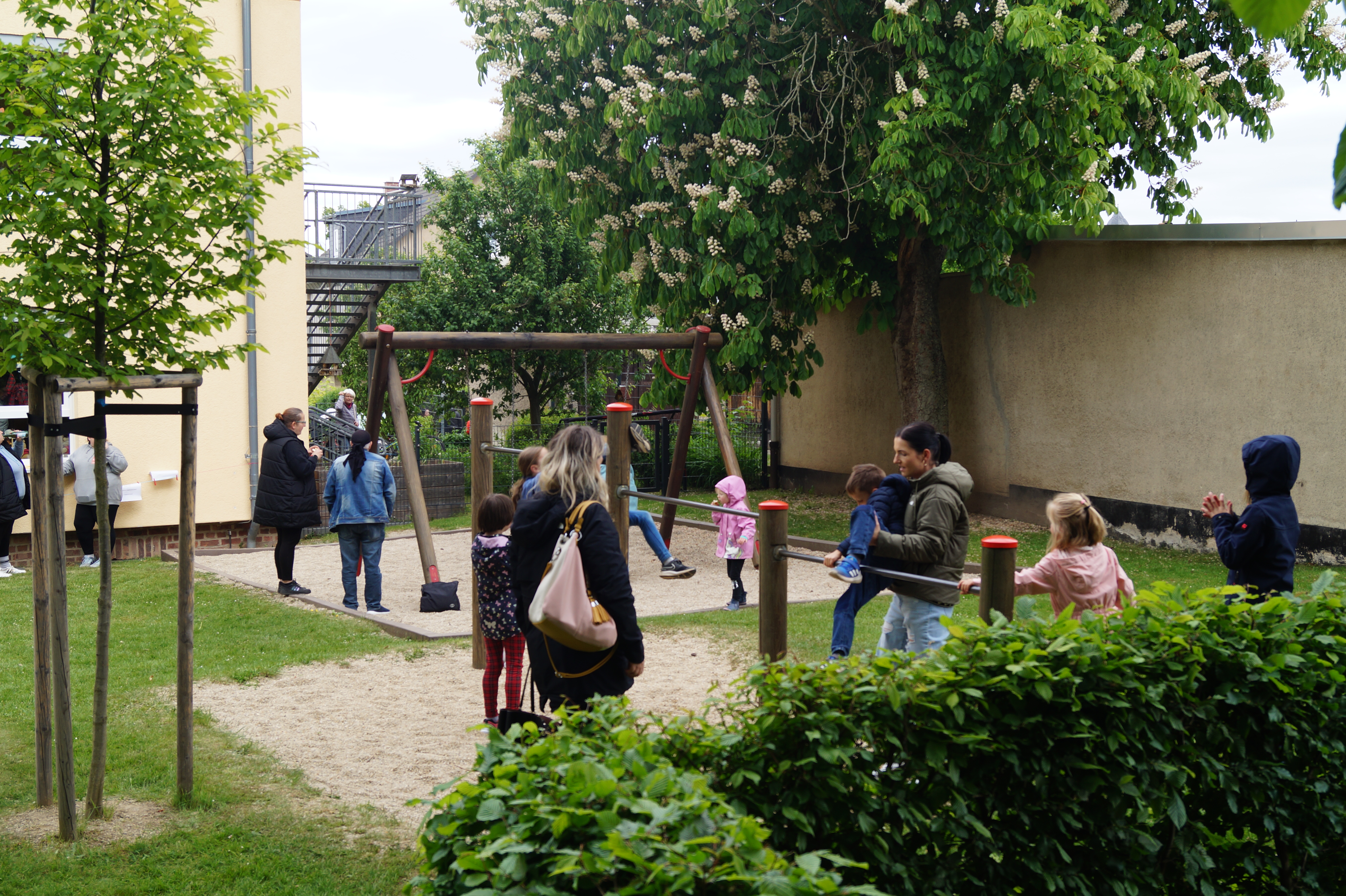 Ein Foto eines kleinen Spielplatzes im Freien, auf dem mehrere Erwachsene und Kinder miteinander spielen und sich unterhalten. Sichtbar sind Schaukeln und Spielgeräte, umgeben von einer grünen Hecke und Bäumen.